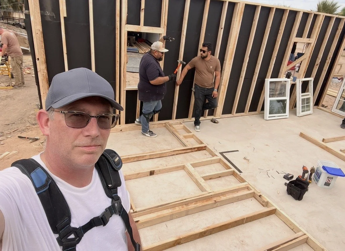 Man wearing glasses and a baseball cap taking a selfie at a construction site with wooden framing, two men talking, and construction tools nearby.