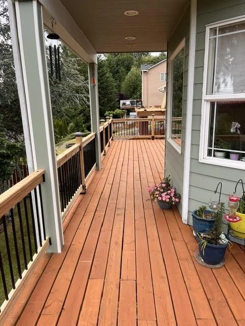 A long wooden porch with a railing, decorated with potted plants and flowers, attached to a house with a window and a view of neighboring houses and trees.