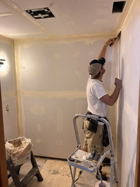 A man painting a wall in a room with a short-piled ladder nearby, and tools and construction materials on the floor.