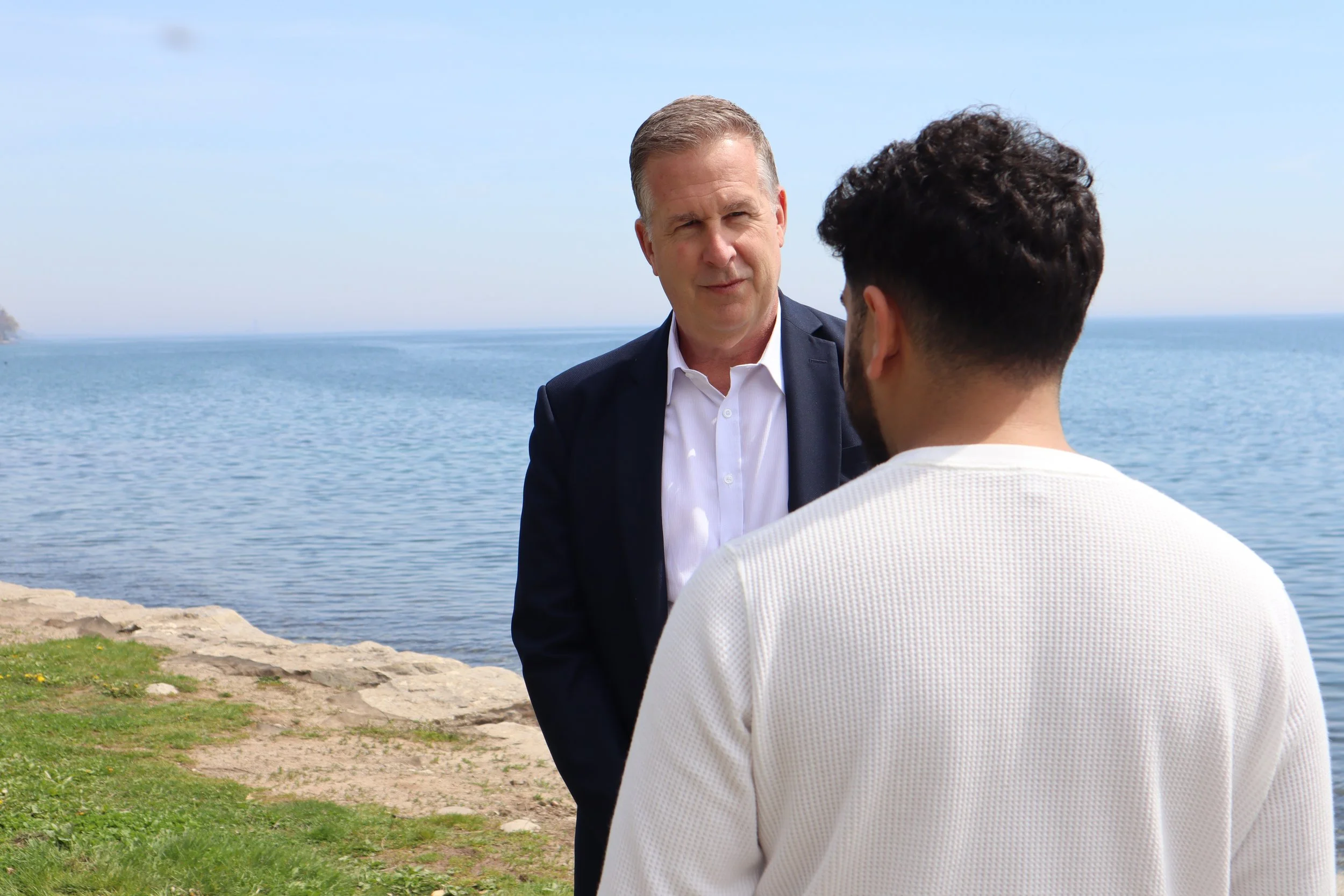 Two men having a conversation by the water on a rocky and grassy shoreline under a clear sky.