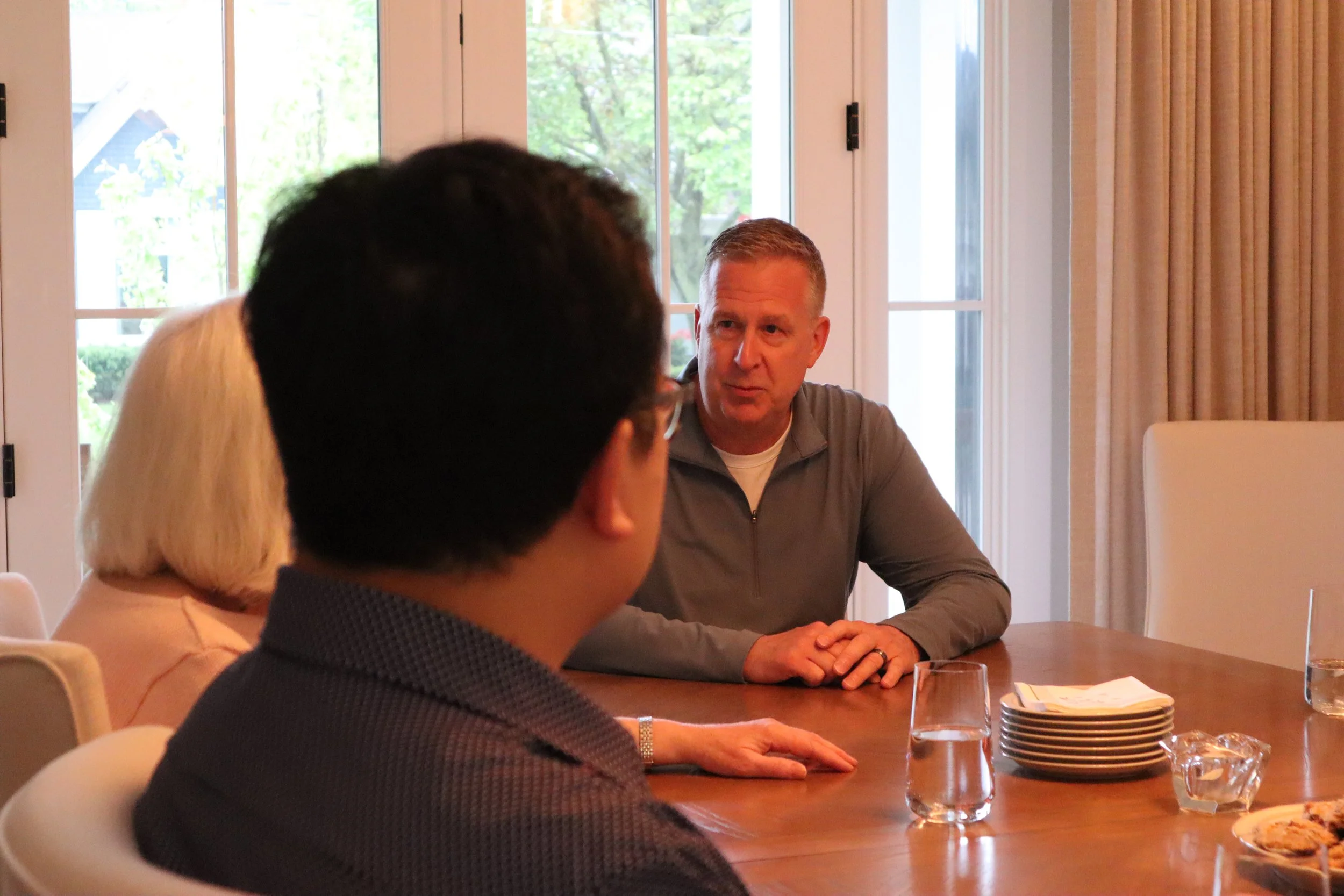 A meeting room with several people seated around a table. A man with short gray hair and a gray shirt is looking at a person with black hair and glasses, who has blonde hair and is wearing a light pink top. The table has glasses of water, plates, and some snacks. Large windows and beige curtains are in the background.