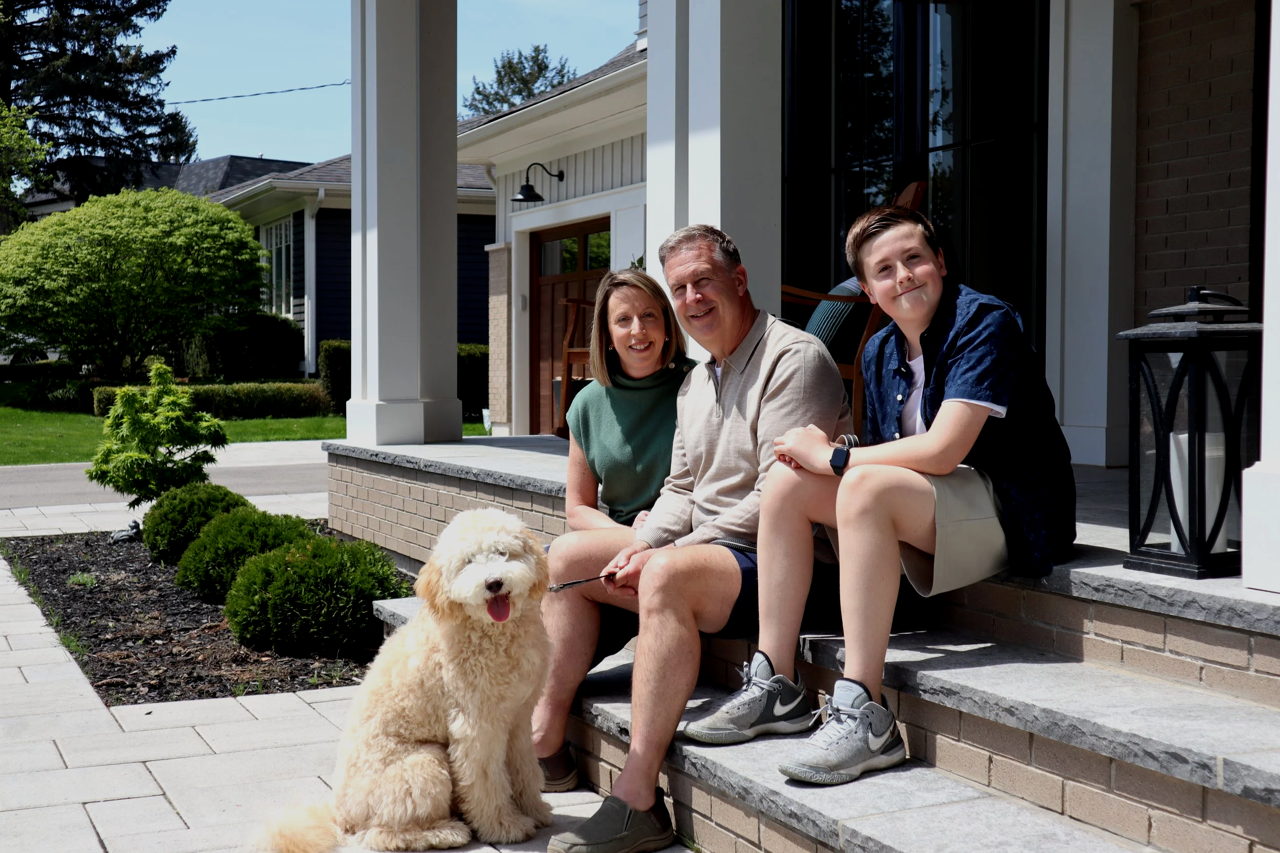 A family of three sitting on the front porch of their house with their dog, smiling at the camera. The porch has granite steps and a small garden with bushes."