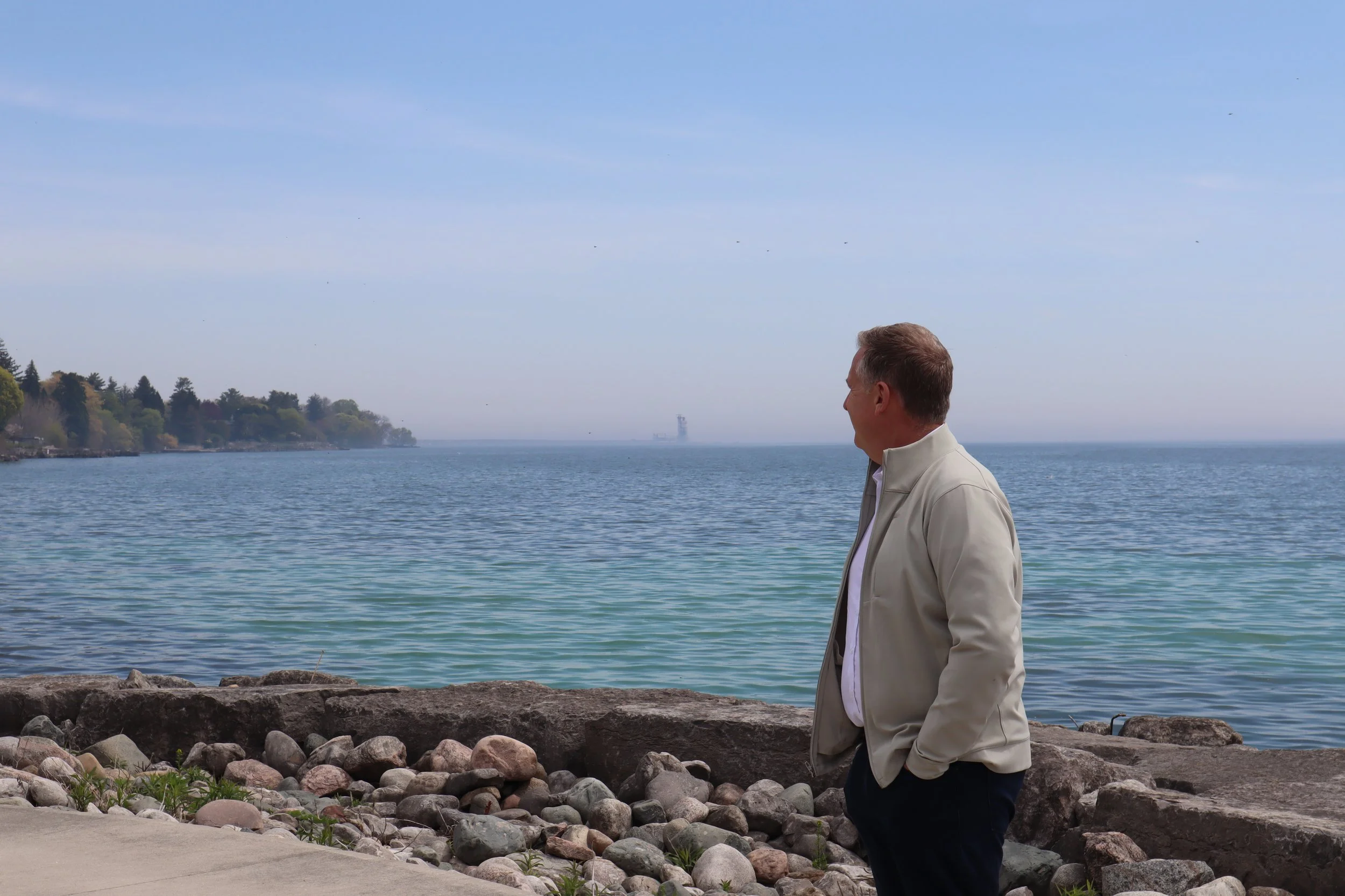 A man with short hair wearing a light-colored jacket and dark pants stands by a rocky shoreline, looking out at the water. The scene includes a calm body of water, distant trees along the coast, and a clear blue sky with a few clouds.