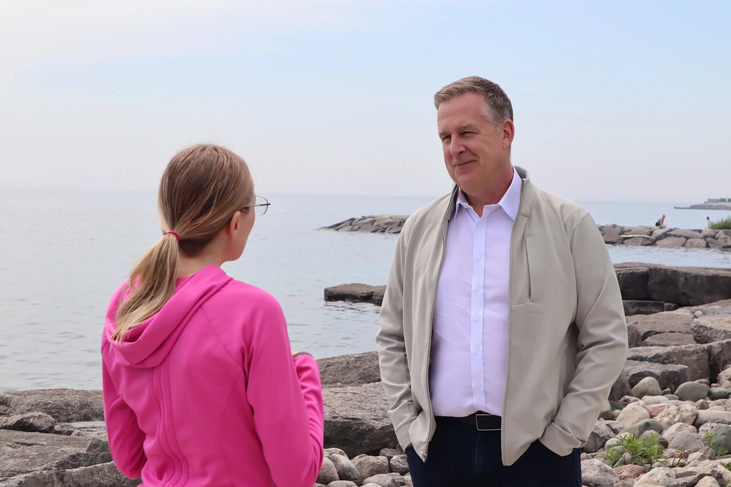 A man with short gray hair, wearing a light beige jacket over a white shirt, standing by rocks at the seaside. He is talking to a young girl with blonde hair tied back with a red hair tie, wearing a bright pink hoodie. The background shows a rocky shoreline, calm ocean, and a cloudy sky.