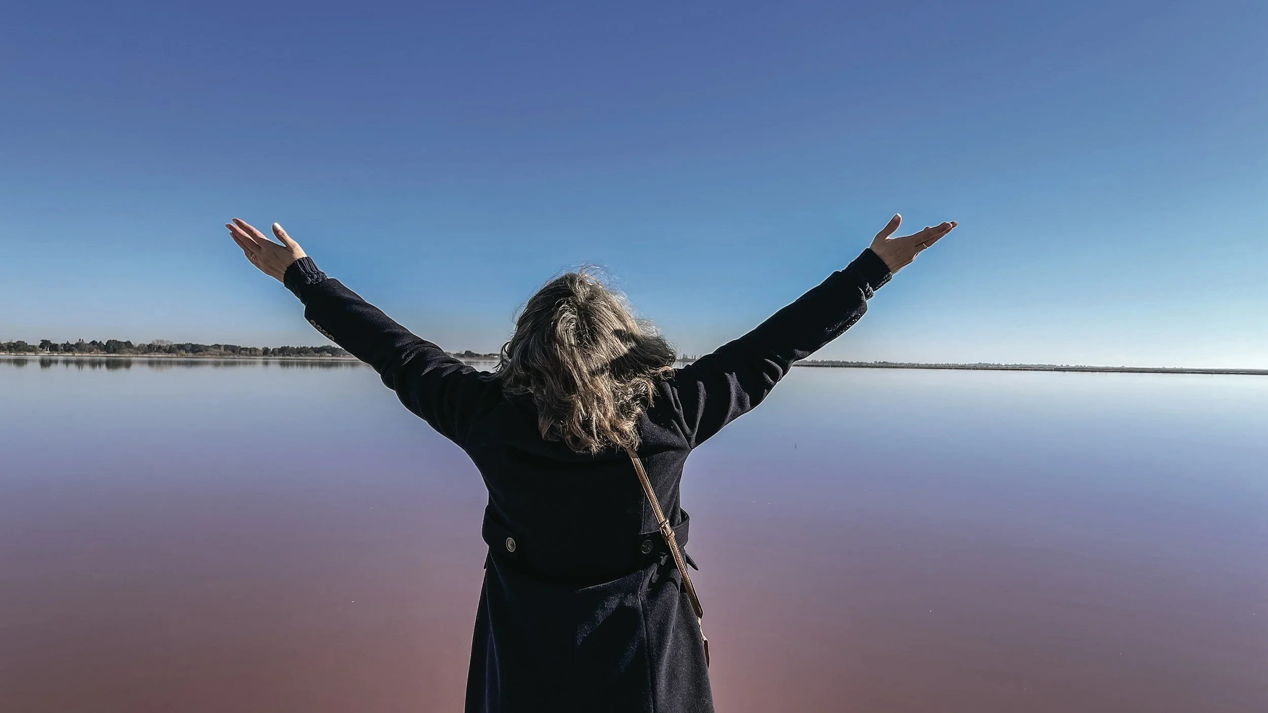 Person with arms raised standing by a calm body of water under a clear blue sky.