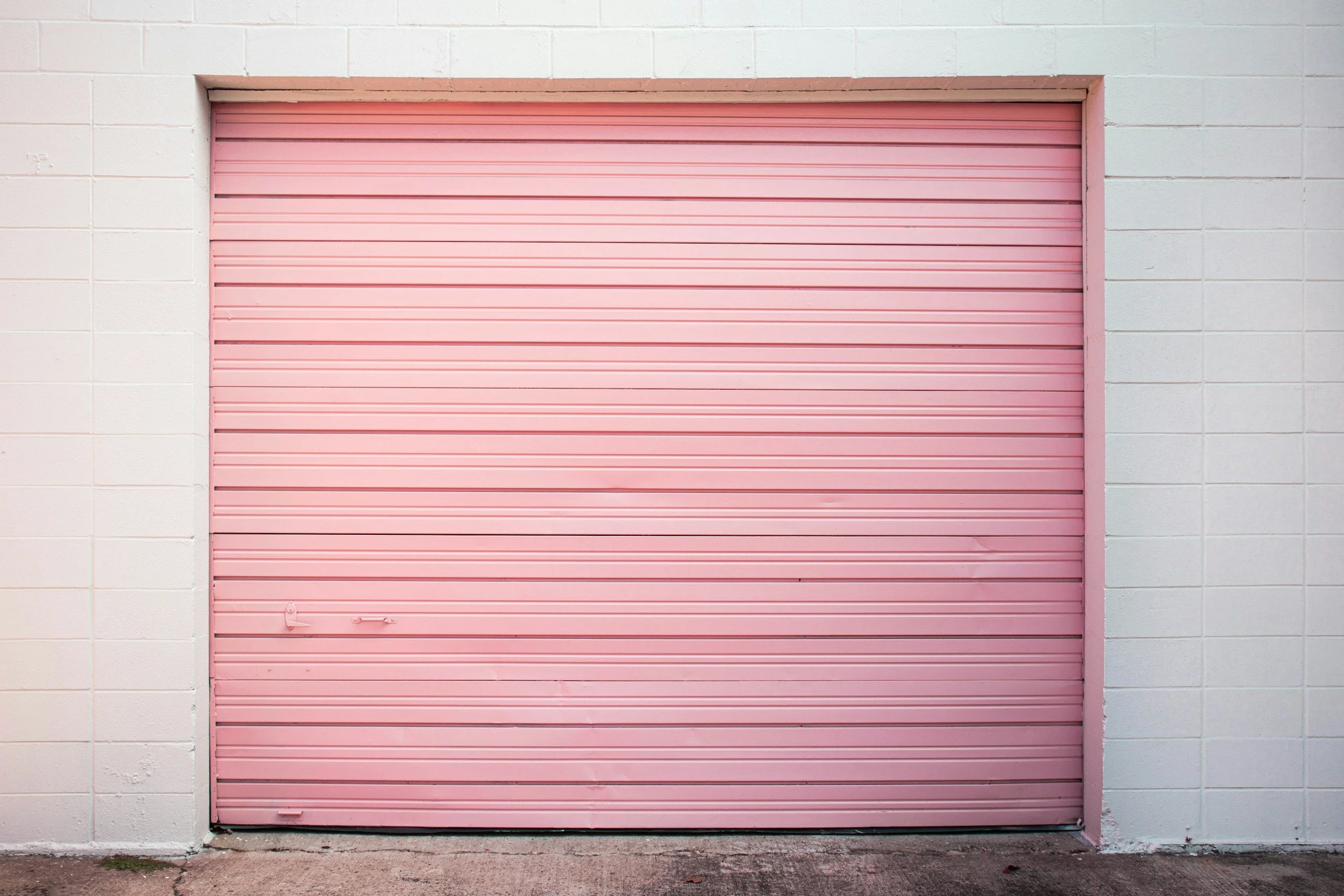 Pink metal garage door with horizontal slats, set in a white concrete block wall, with a concrete ground in front.