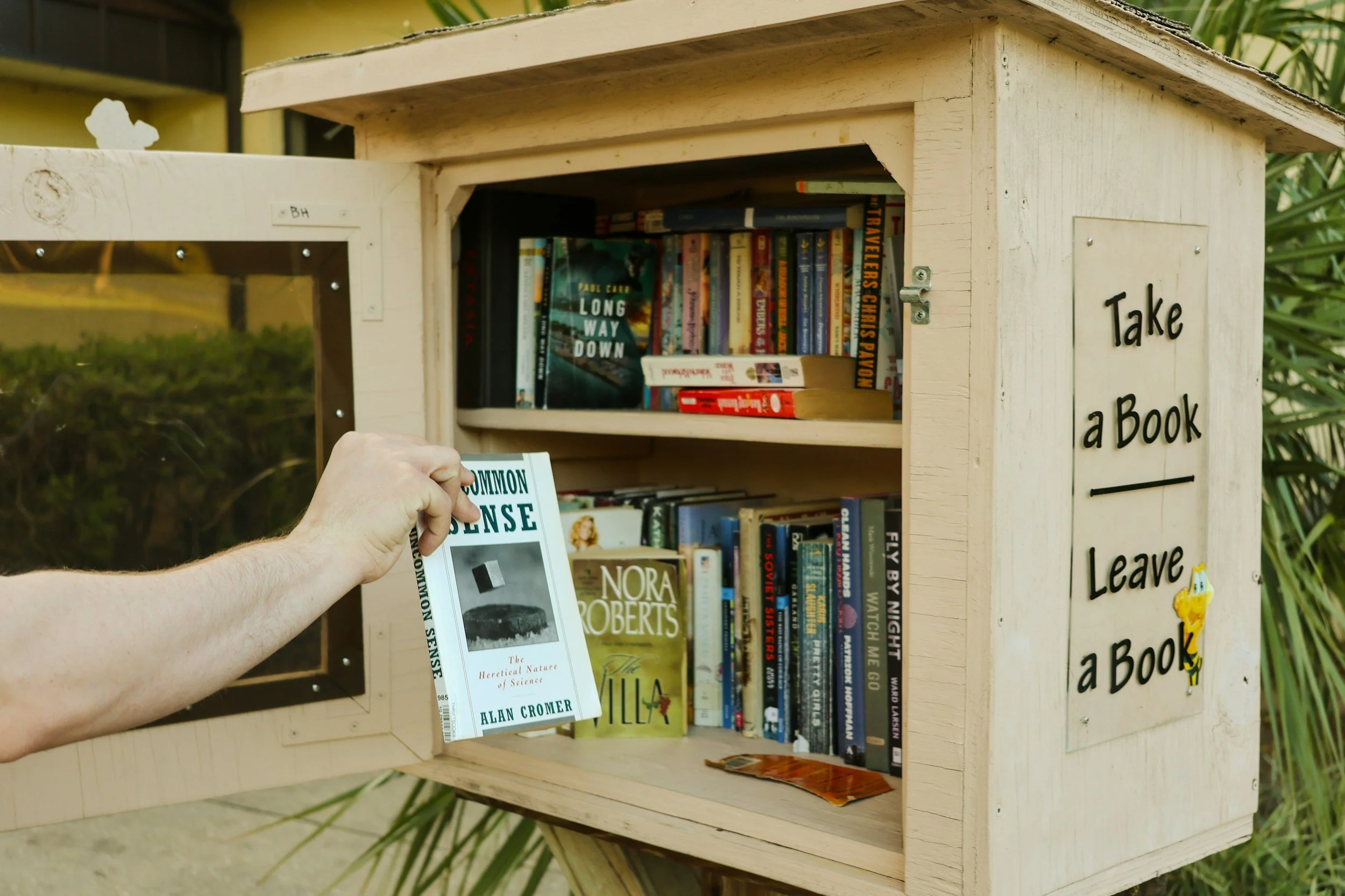 A person is placing a book into a small, wooden community book exchange box labeled "Take a Book, Leave a Book." The box contains several books, including titles like "Long Way Down" and "Nora Roberts."