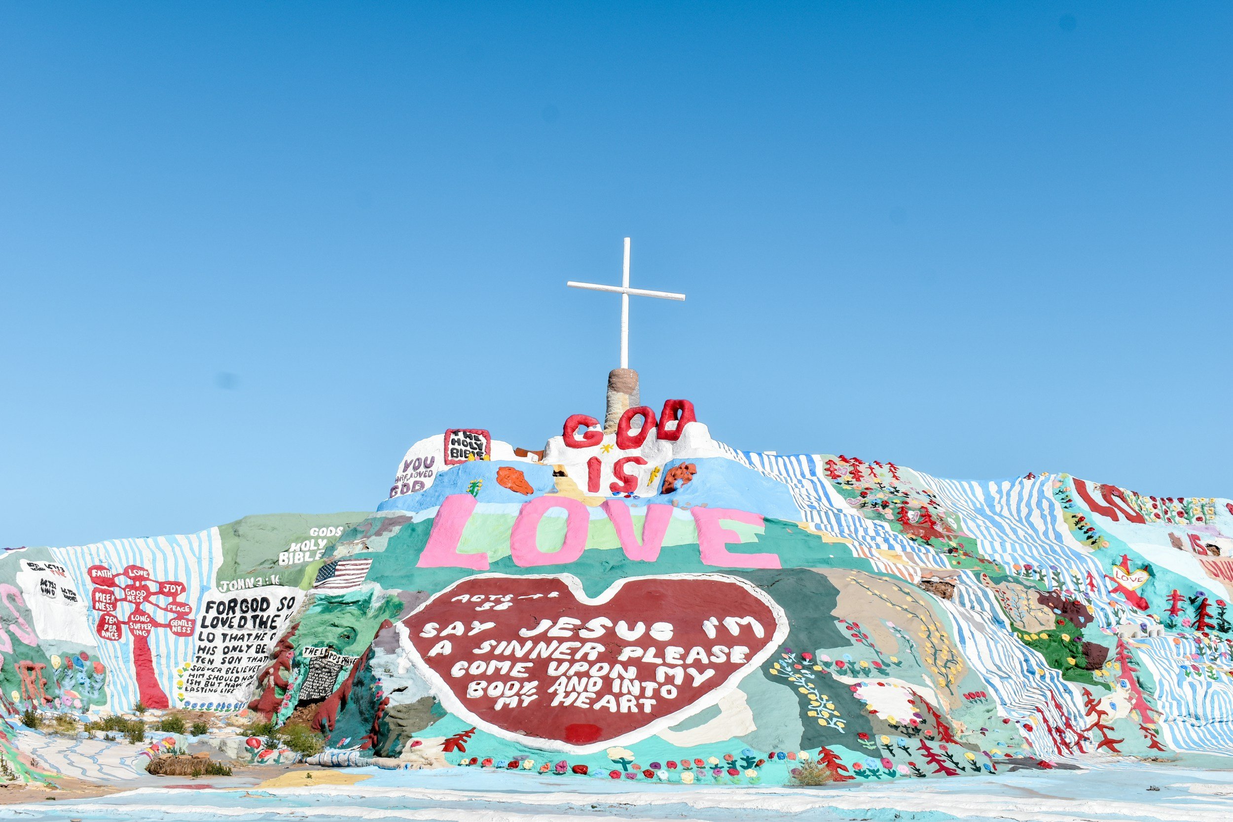 Colorful hillside with religious and inspirational messages, drawings, and symbols including a cross at the top against a clear blue sky.