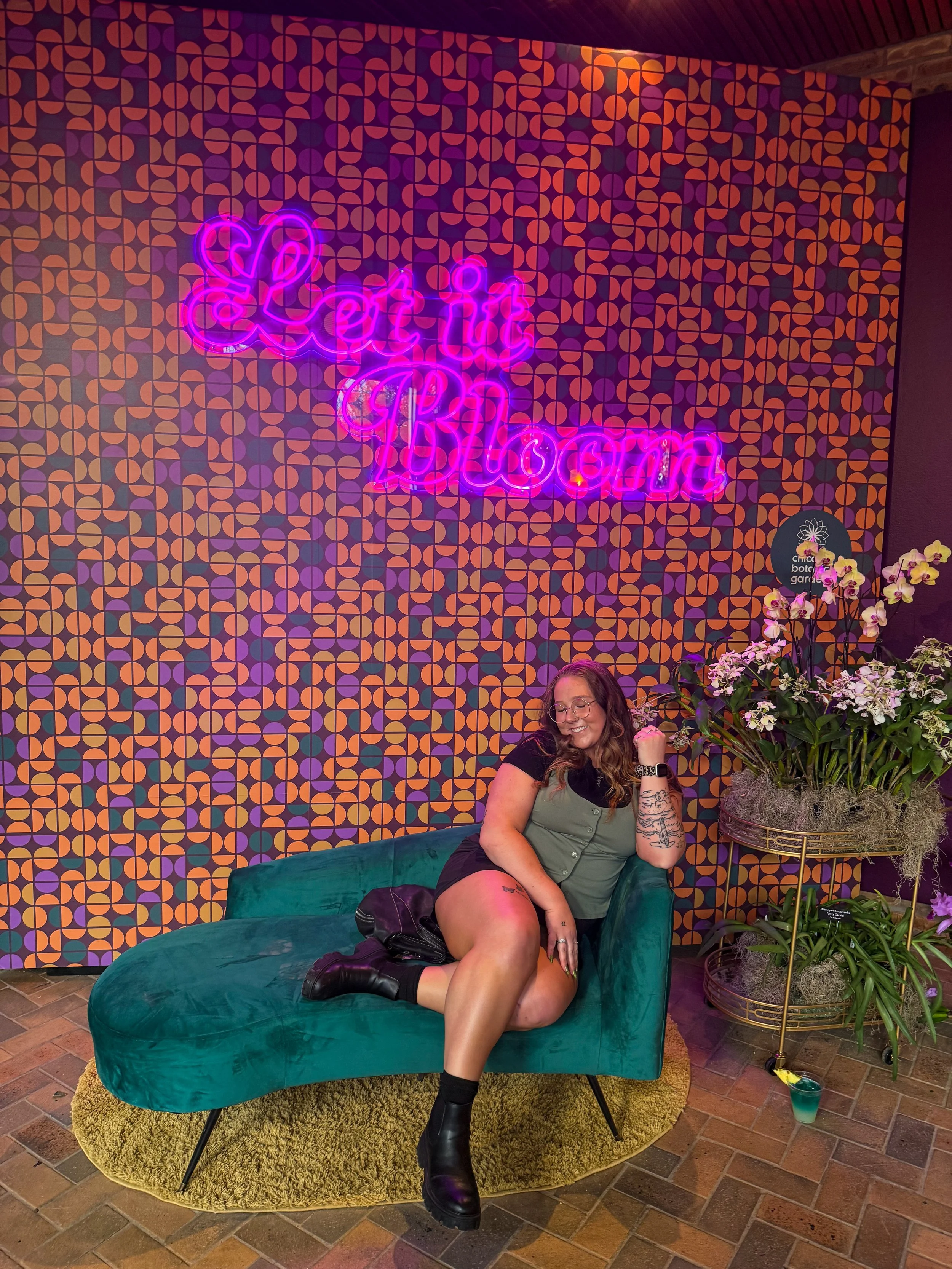 A woman with glasses and tattoos sitting on a green velvet sofa in front of a patterned purple and orange wall with a neon sign that reads 'Let it Bloom.' She is smiling, with her left hand near her face. There is a floral arrangement in a basket on a gold stand to her right, and a drink on the floor beside the sofa.