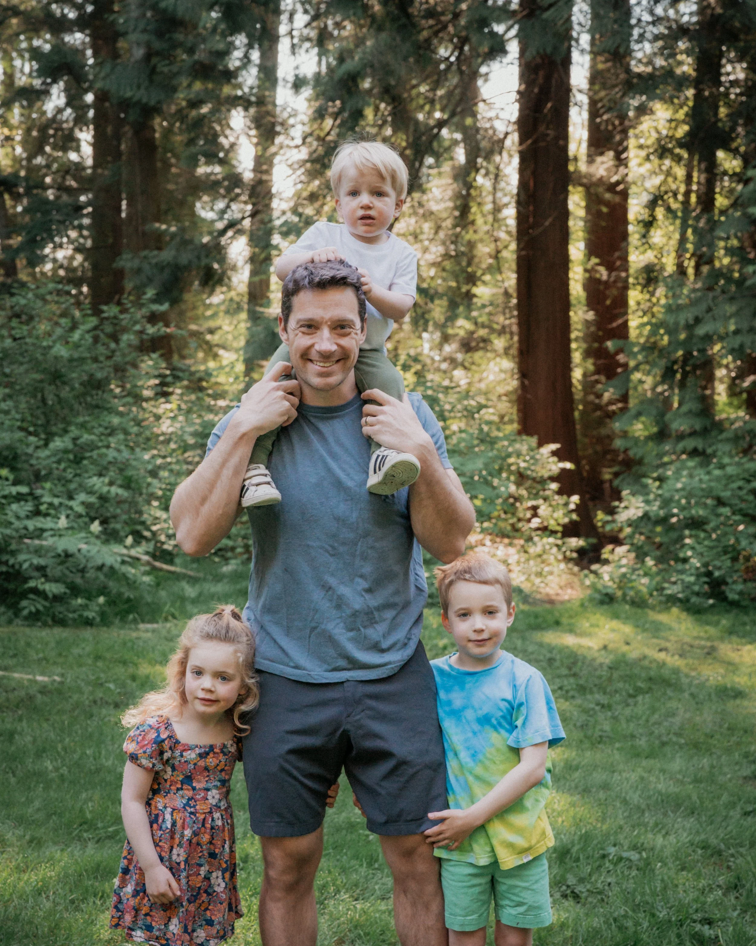 A man with three children in a forest. The man is smiling, wearing a gray t-shirt and shorts, and has a child on his shoulders. The children are two boys and a girl, standing on a grassy area with tall trees in the background. The girl is wearing a f