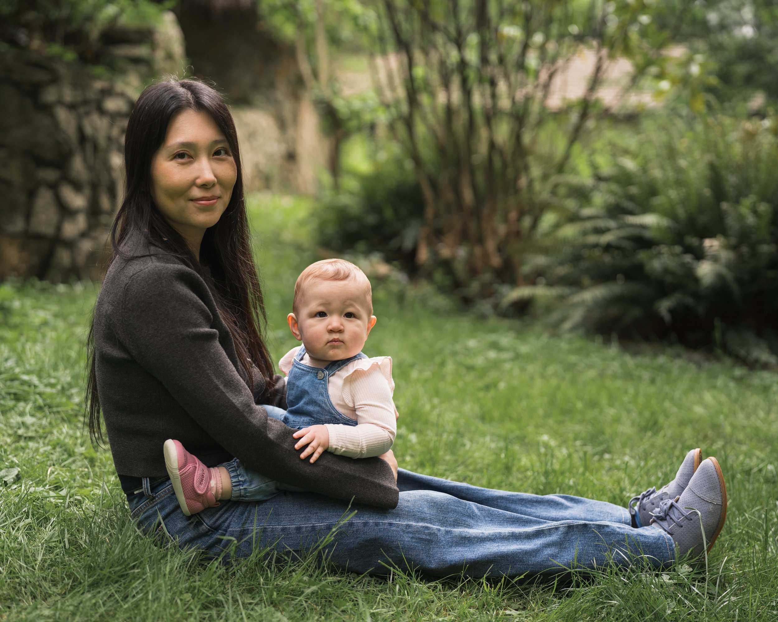 A woman with long dark hair sitting on the grass in a park, holding a baby, both looking at the camera. The woman is wearing a dark jacket and jeans, and the baby has short curly hair, a beige top, and denim overalls. The background features green tr