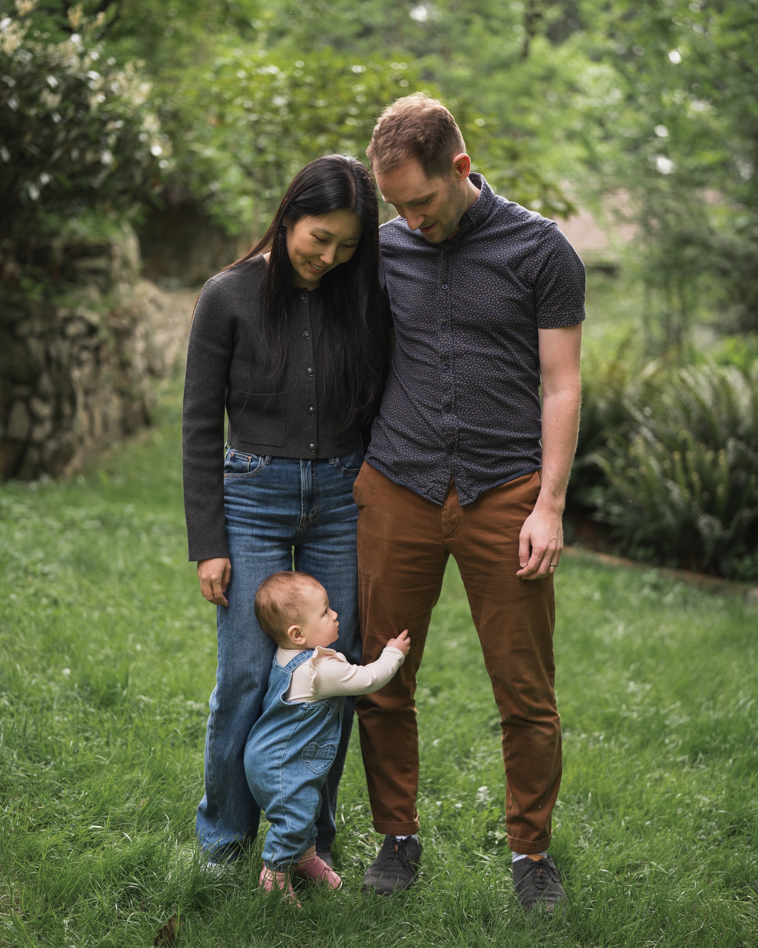 A woman, a man, and a young child standing together outdoors in a grassy area with trees. The woman and man are looking down, smiling, while the child, hugging the man's leg, looks up at him.