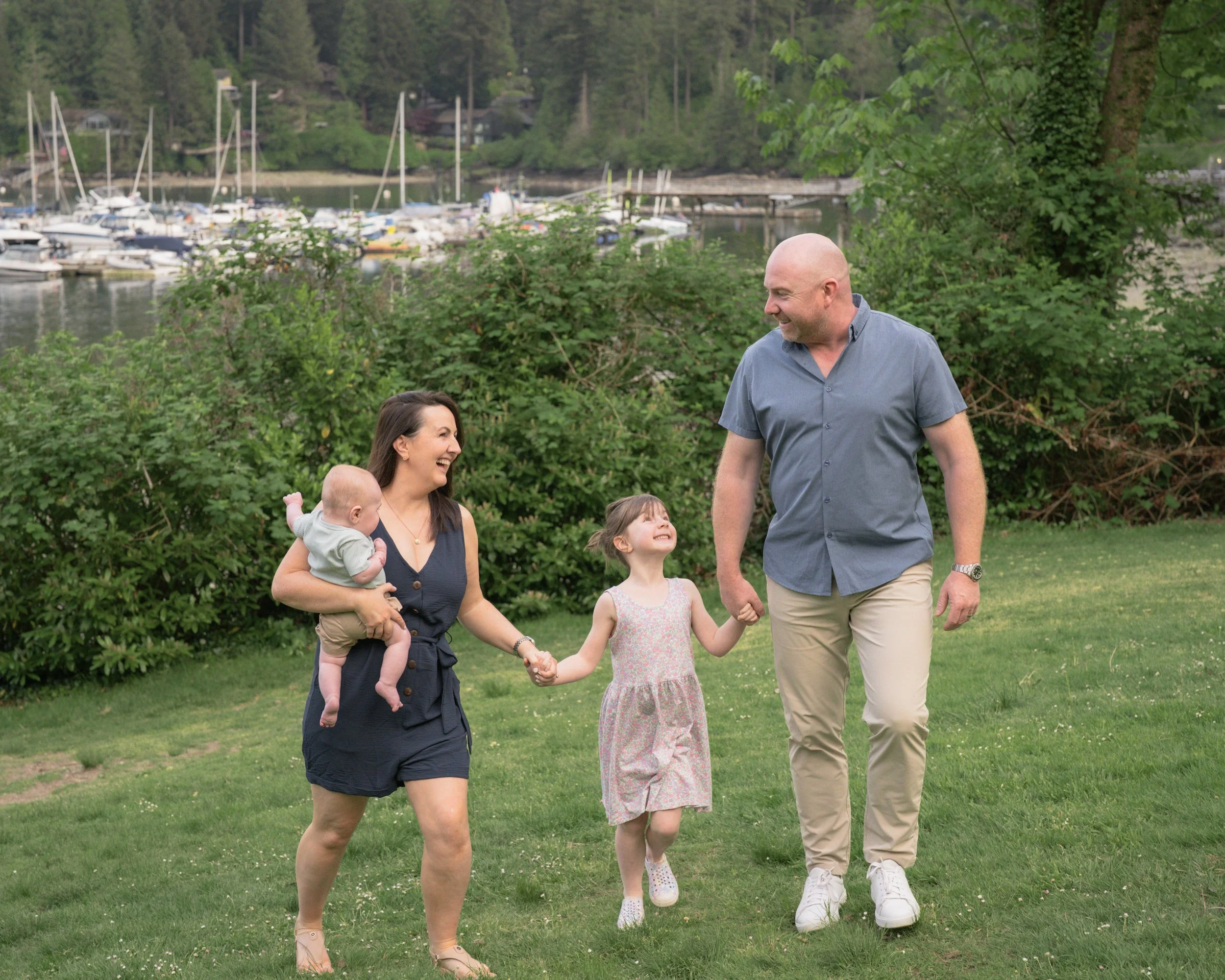 A family of four, including a woman holding a baby, a young girl, and a bald man, are walking and smiling outdoors near a marina with boats and trees in the background.