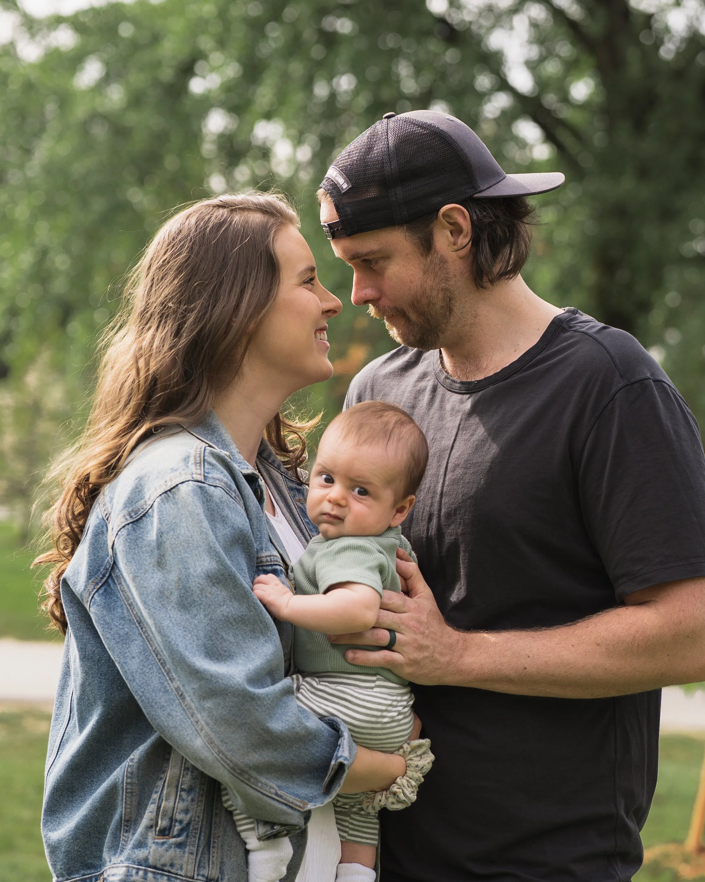 A couple is standing close outdoors with two children, a baby and a girl, smiling at each other amid green trees.