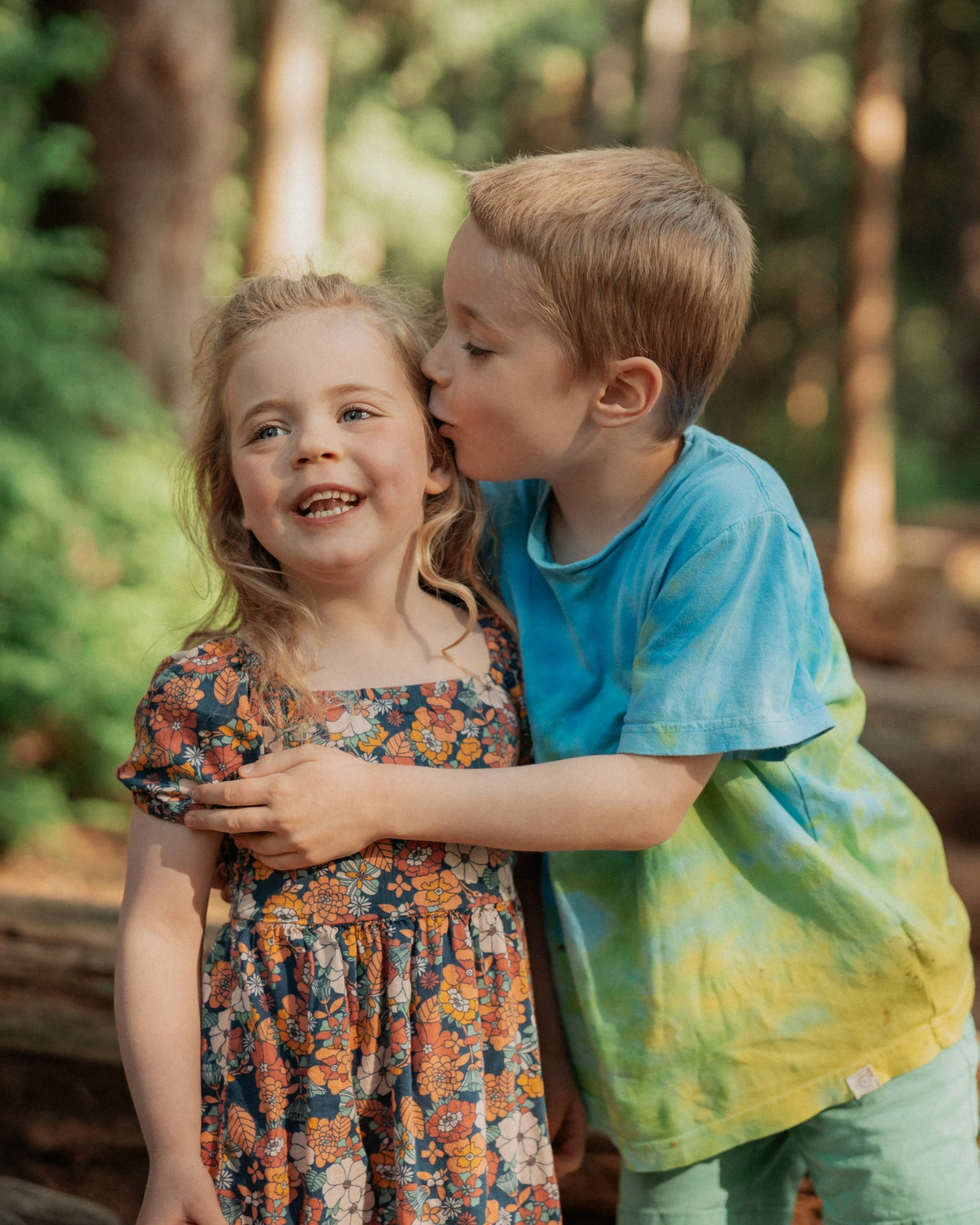 A young girl with curly blonde hair wearing a floral dress being hugged and kissed on the cheek by a boy with short light brown hair wearing a blue t-shirt. They are outdoors in a forested area with blurred trees in the background.