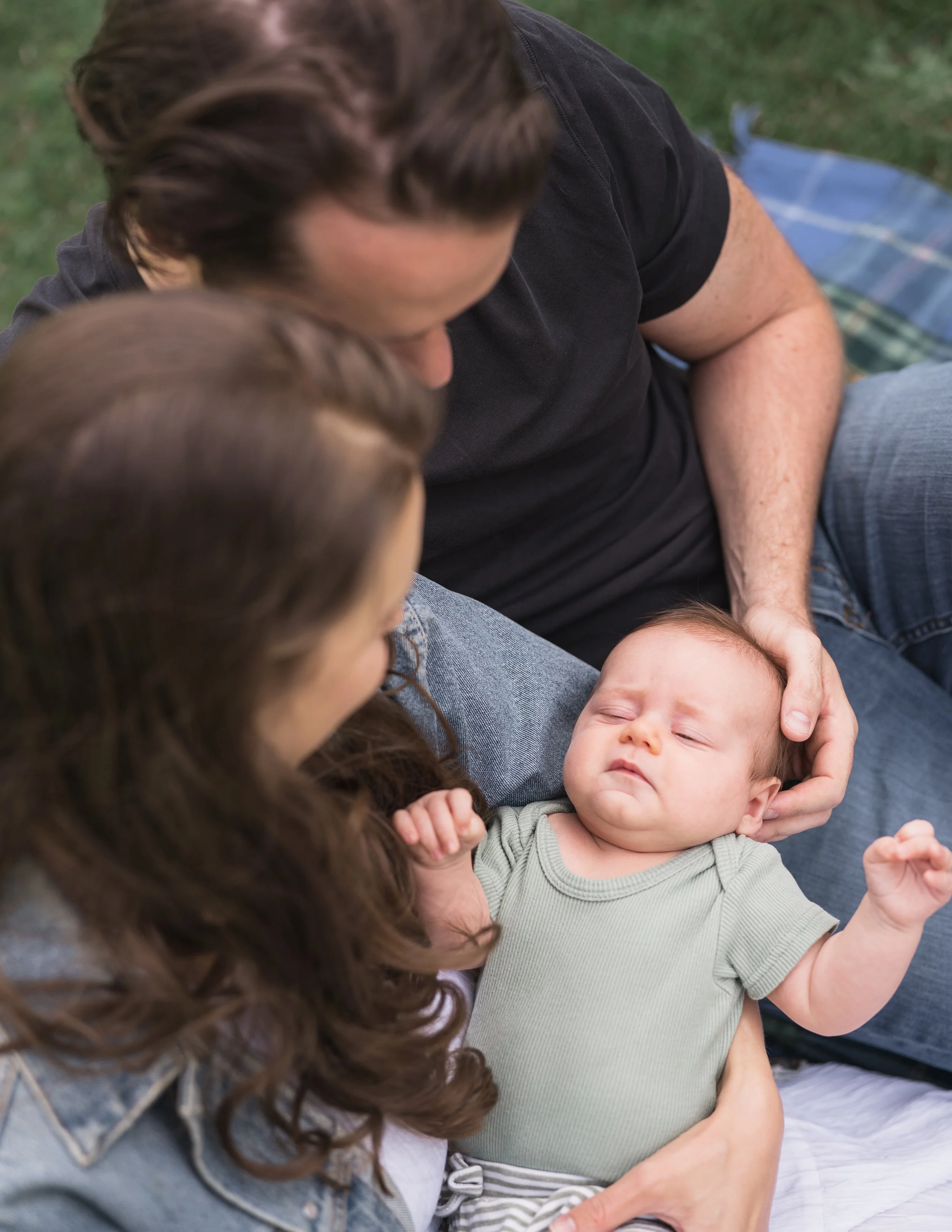 A woman, a man, and a baby are lying on the grass, with their foreheads touching, creating an intimate and affectionate moment outdoors.