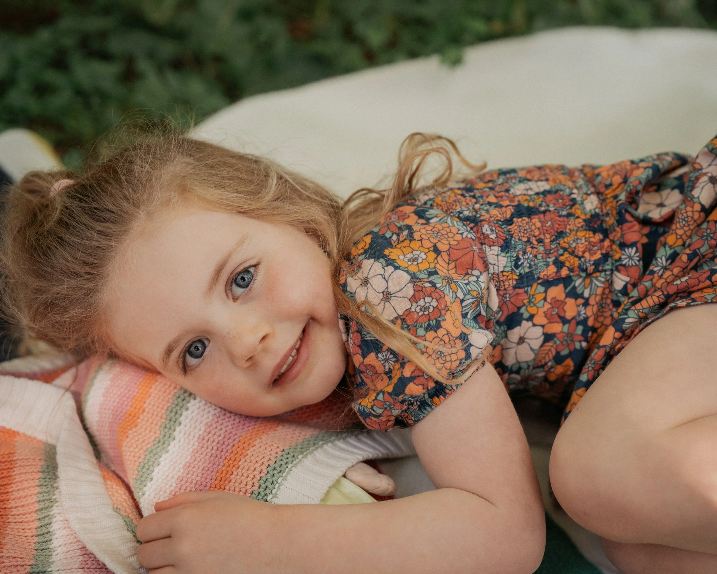 Close-up of a young girl with curly red hair and blue eyes lying on a colorful striped towel, wearing a floral dress, smiling softly.