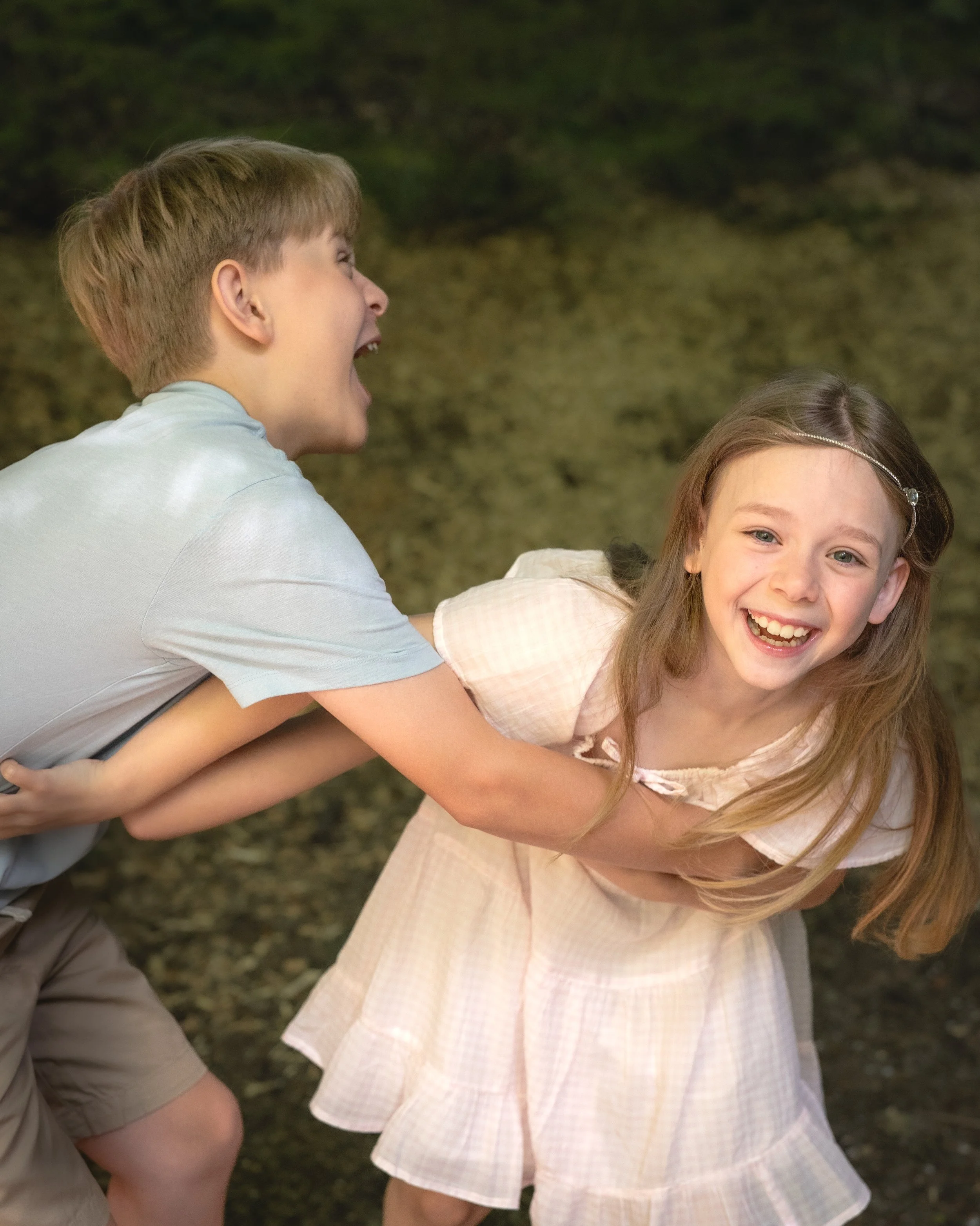 Two children, a boy and a girl, are outdoors on a grassy area, playfully leaning on each other and smiling. The boy has short light brown hair and is wearing a white T-shirt. The girl has long light brown hair with a headband and is wearing a light-c