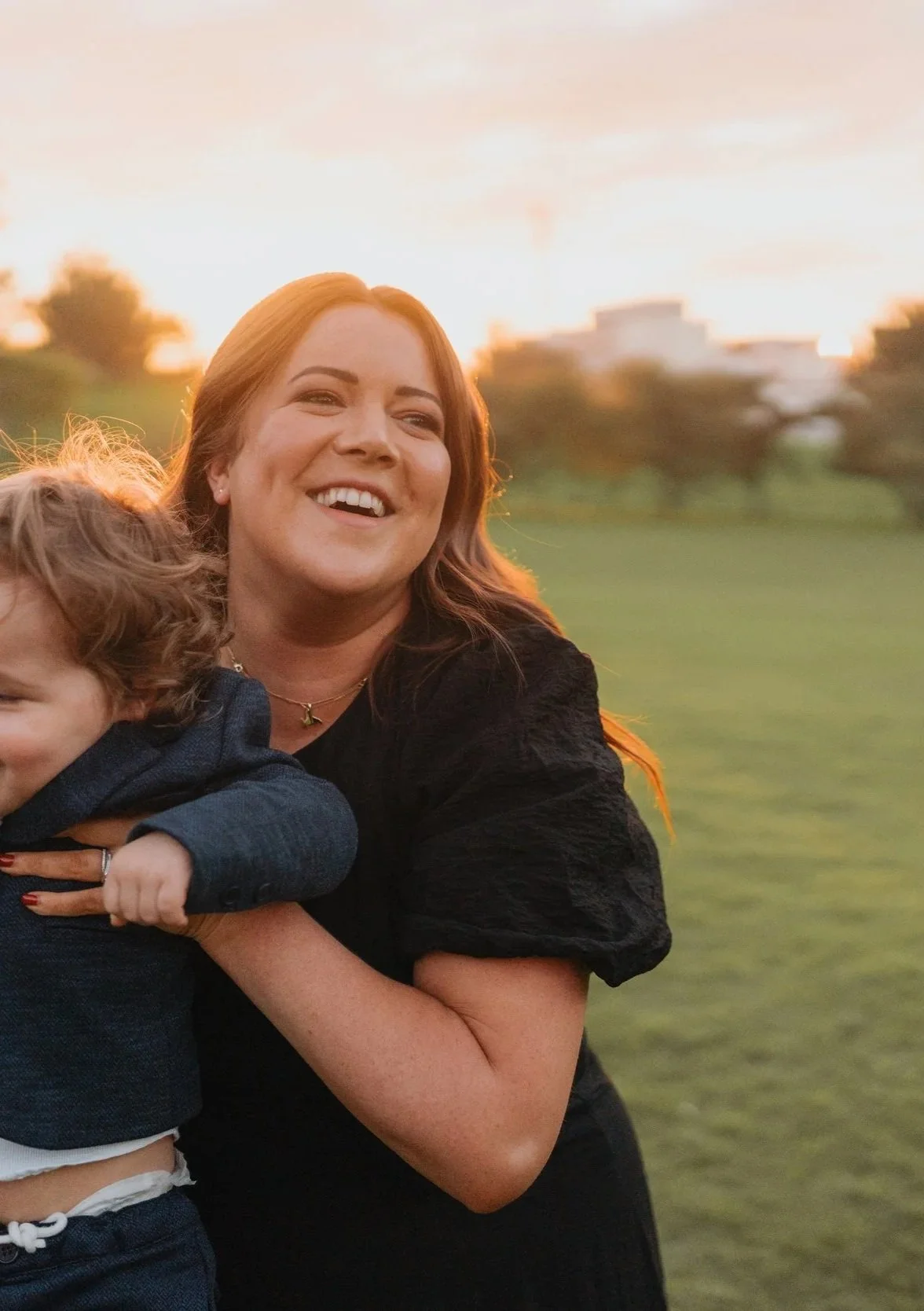 A woman with long red hair smiling and holding a child in an outdoor setting during sunset, with a blurred background of grass and buildings.
