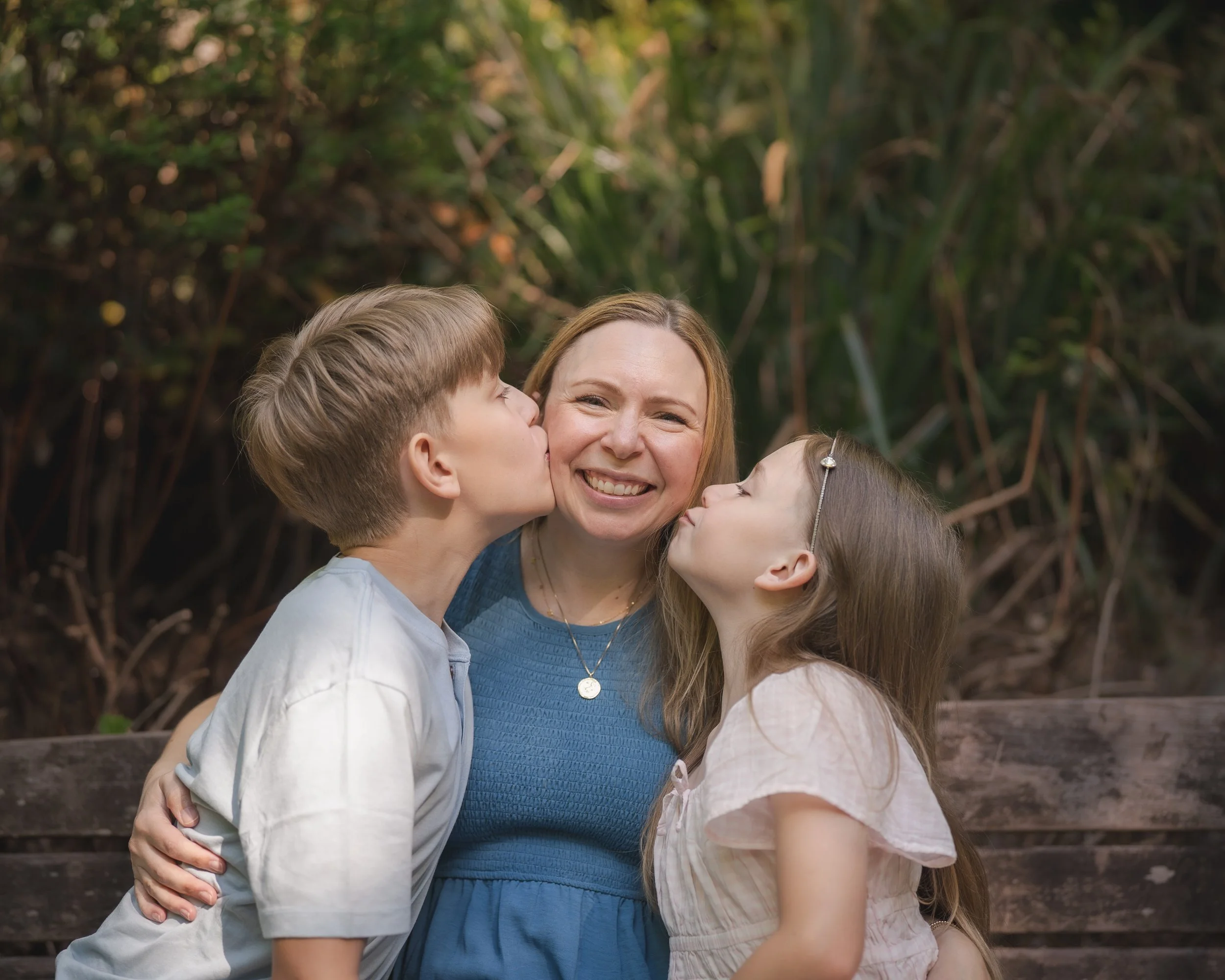 A woman with long hair smiling as she is kissed on the cheek by two children, a boy and a girl, in front of a background of dense green foliage.