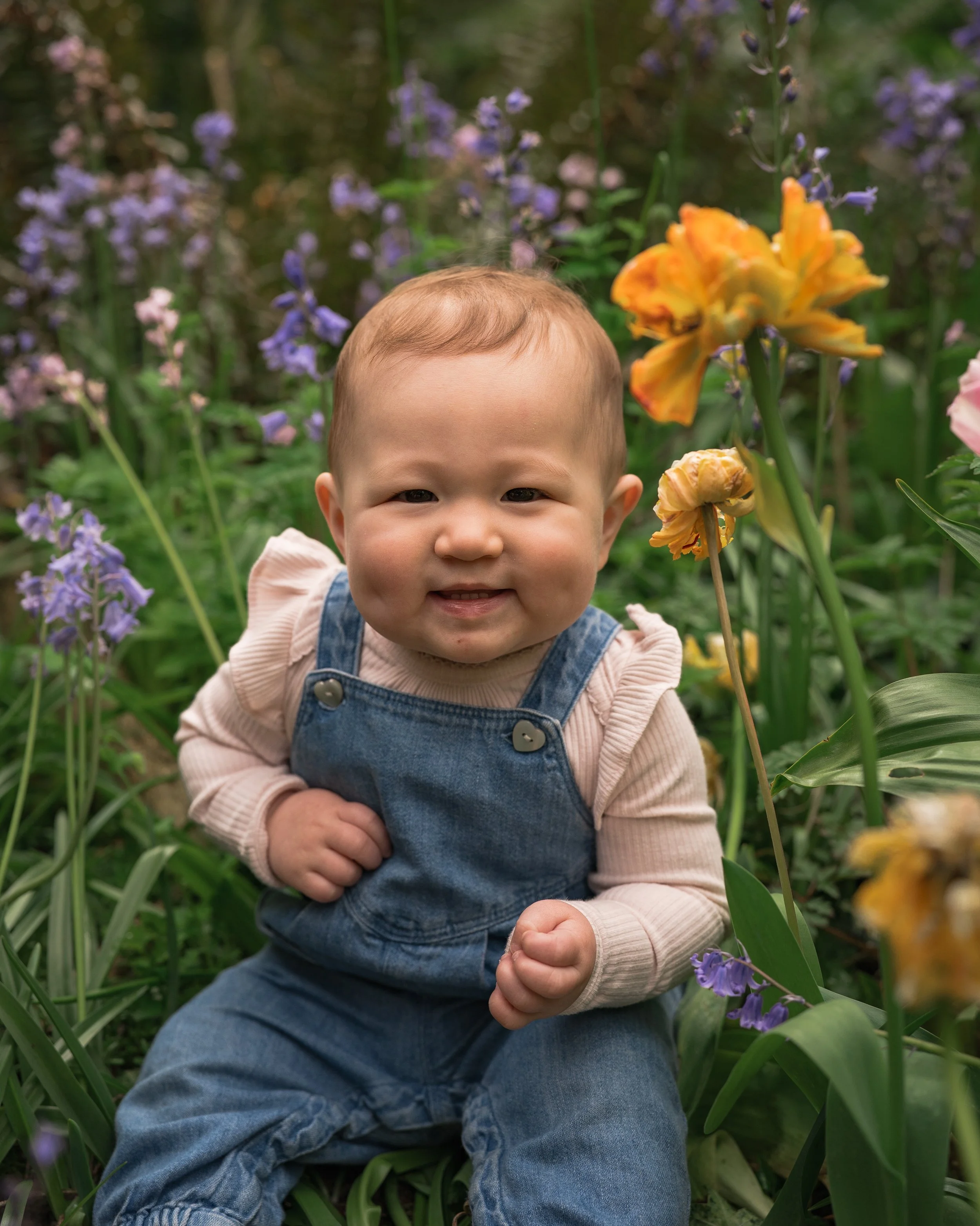 A smiling toddler with light skin and short hair, wearing denim overalls and a long-sleeve shirt, sitting among purple and yellow flowers in a garden.