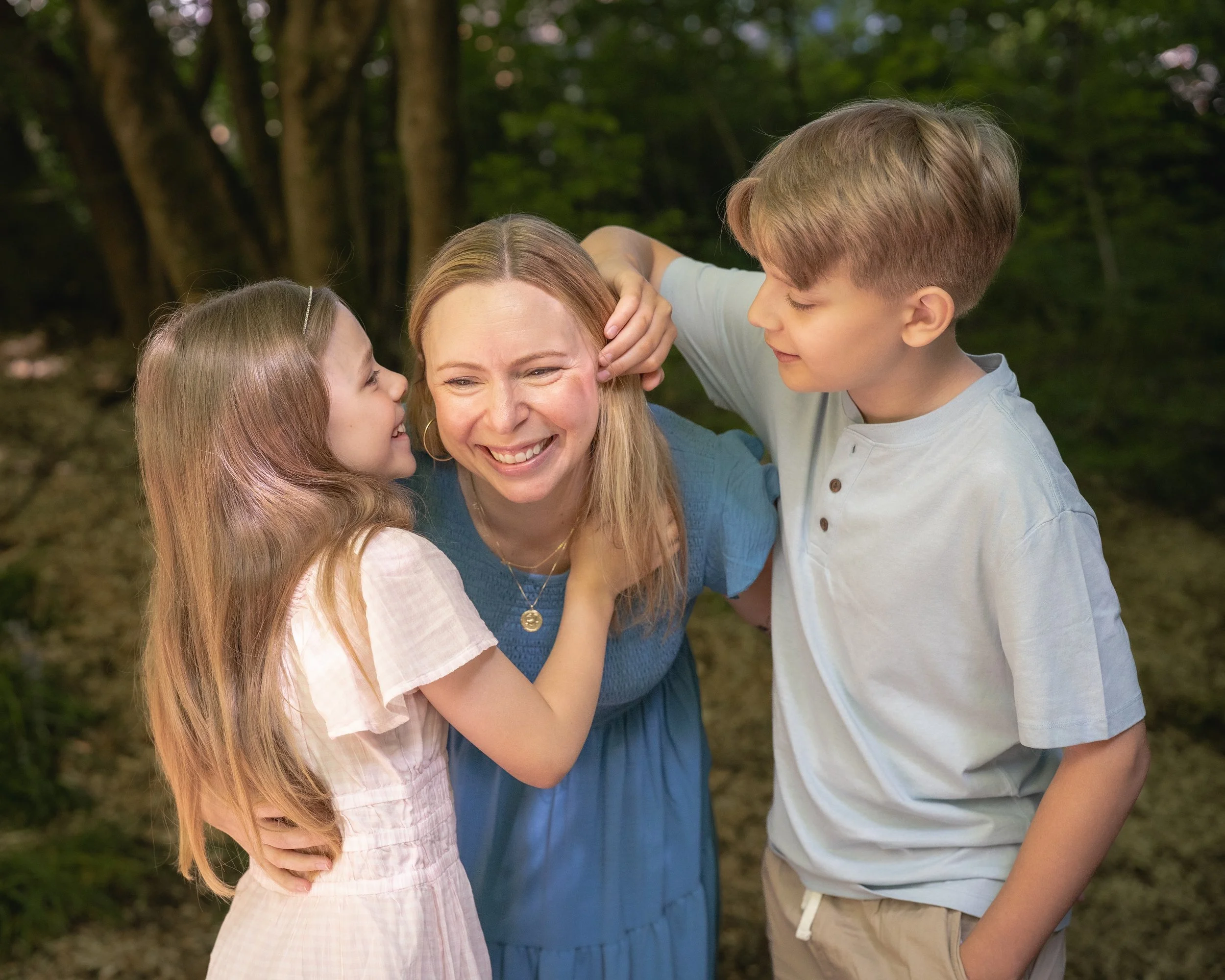 A woman and two children are smiling and hugging outdoors in a wooded area.