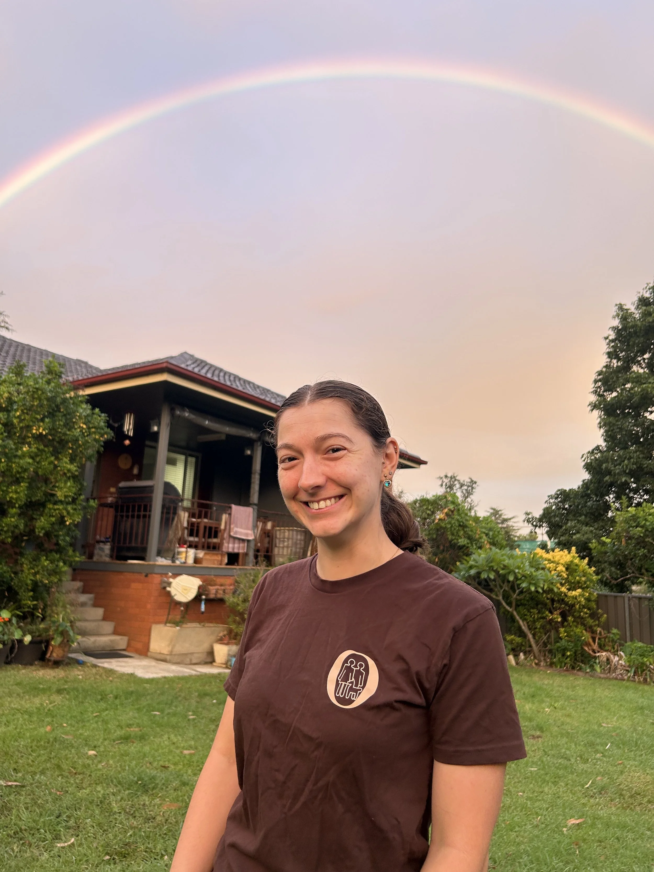 A young woman with dark hair tied back, wearing a brown T-shirt with a logo of two cats, standing outdoors in a backyard with a house, trees, and a rainbow in the sky during sunset.