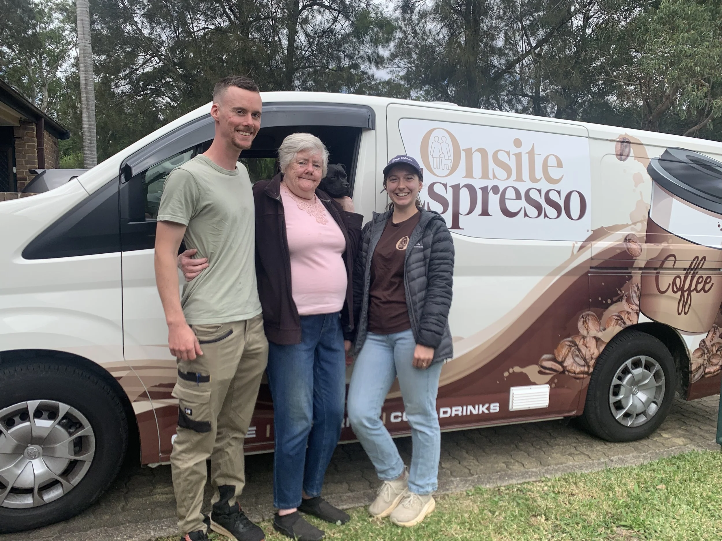 Three people standing together in front of a coffee-themed vehicle, smiling outdoors. One man, a woman in a pink shirt, and another woman in a jacket.