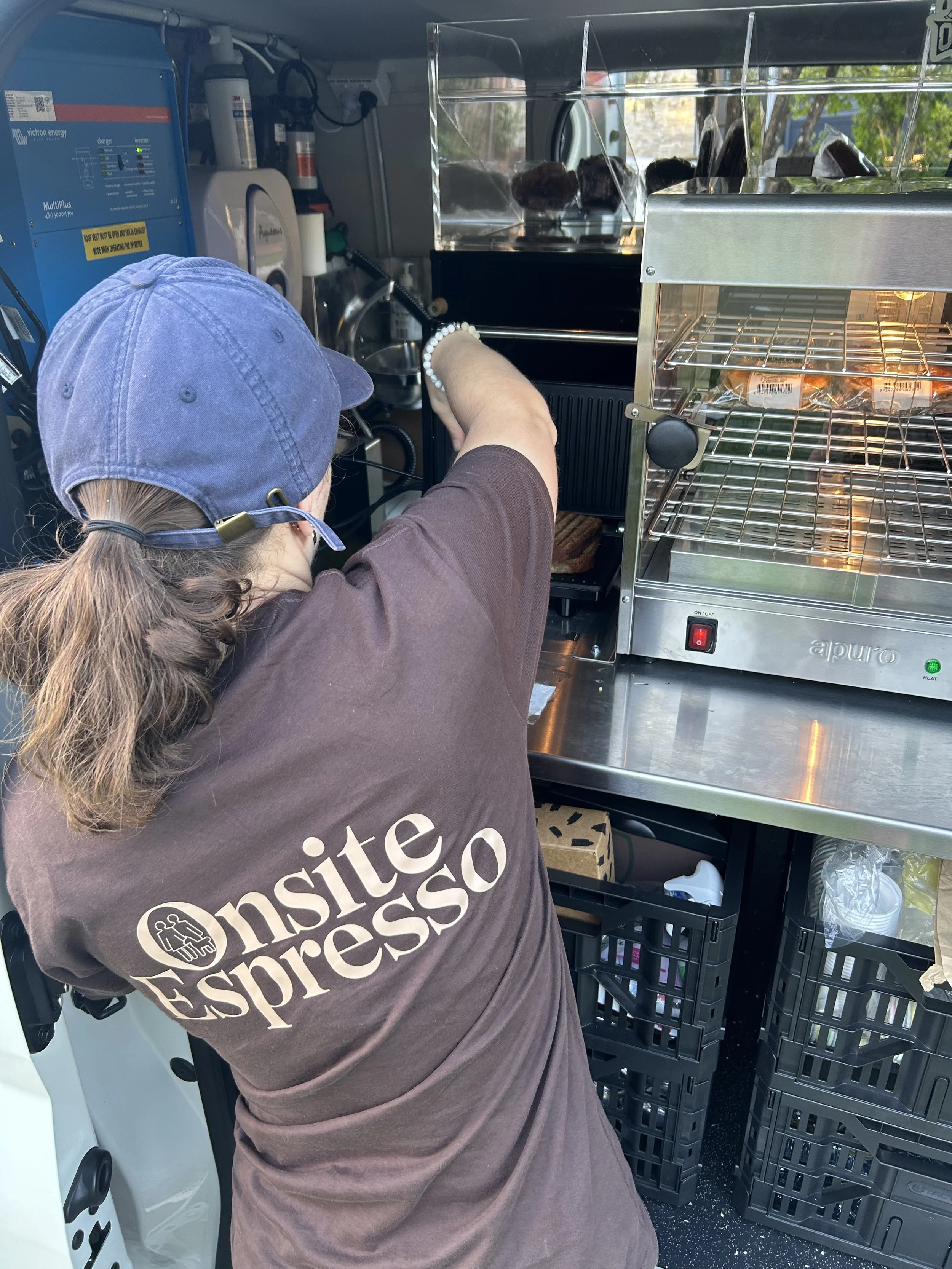 Person wearing a blue cap and a brown shirt with the words 'on site to impress' is reaching into a food truck or cart, with various food containers and equipment visible inside.