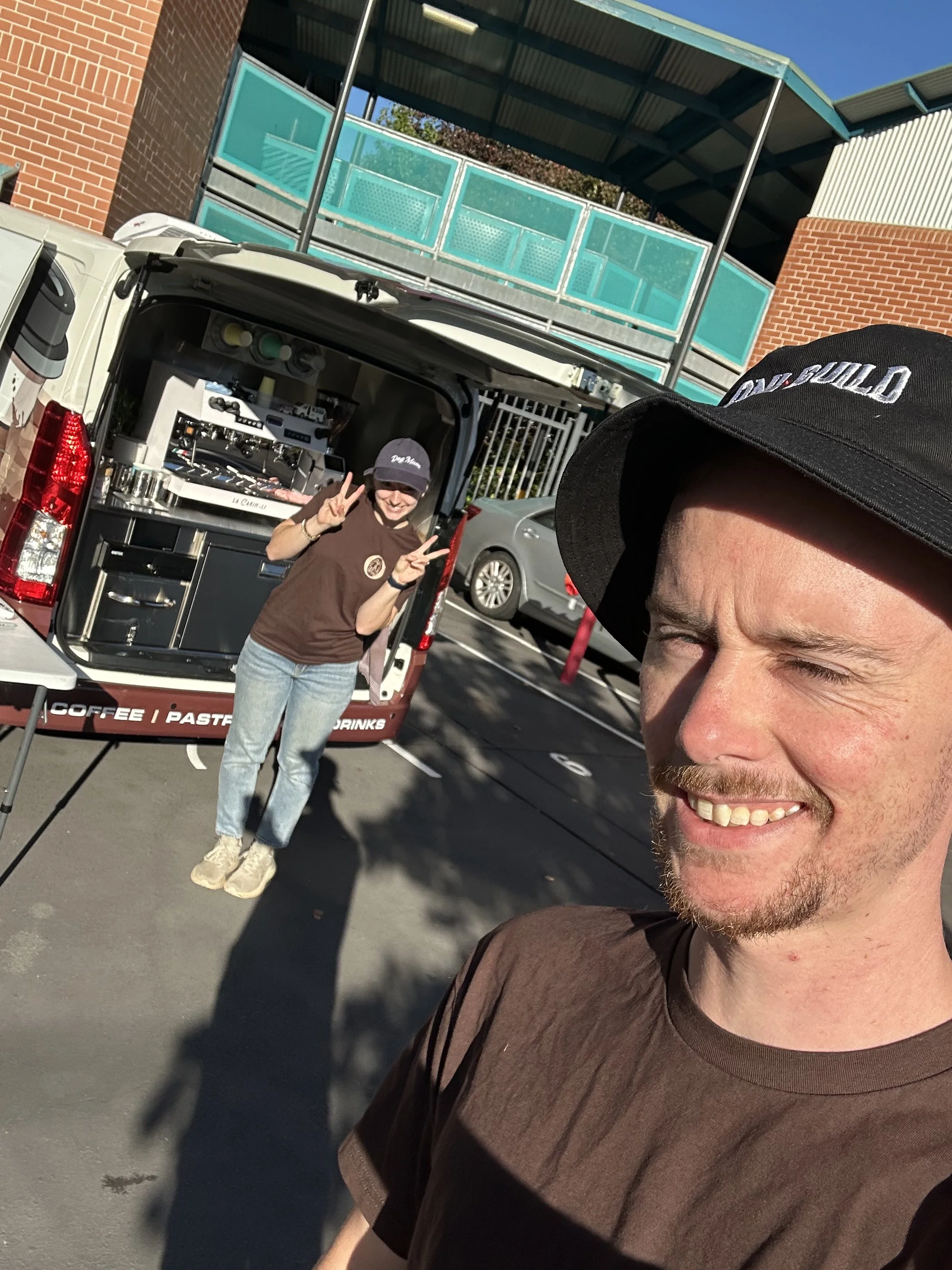 A man taking a selfie smiling with a woman standing happily beside a food truck serving coffee, pastries, and drinks in a parking lot near a building with brick and glass features, sunny weather.