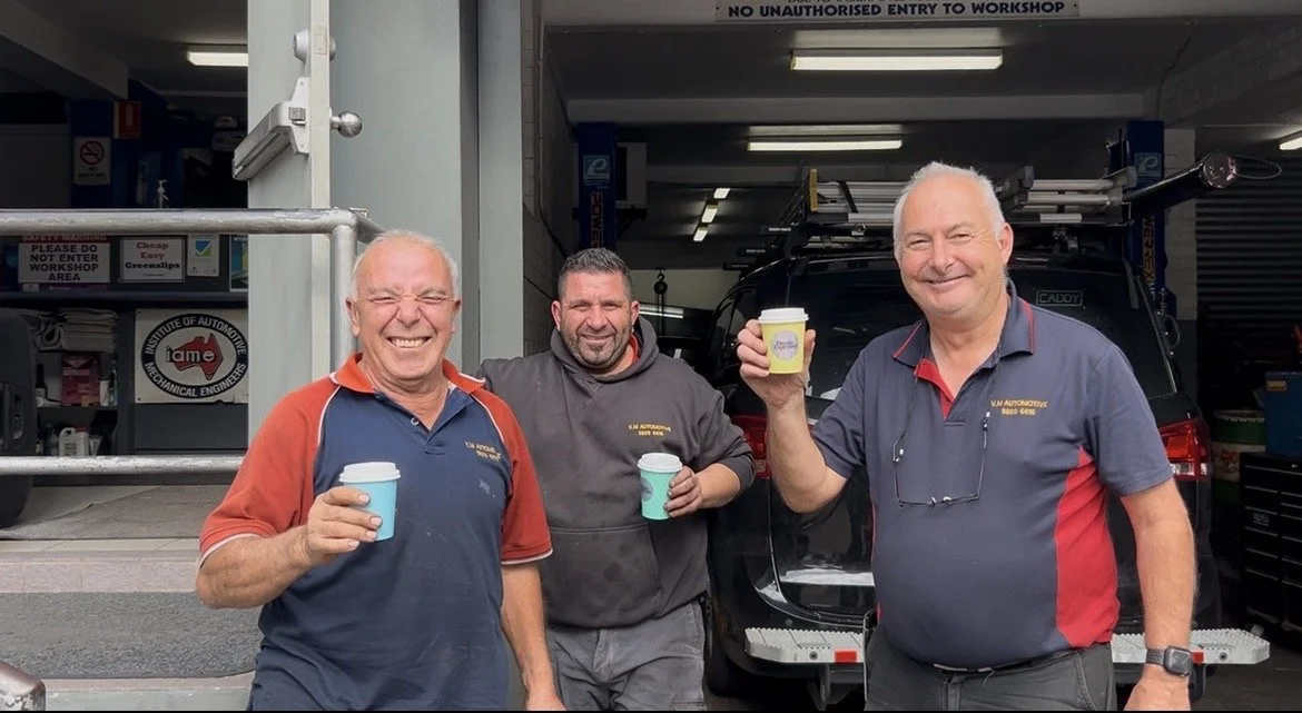 Three smiling men standing outdoors in front of a garage, each holding a coffee cup. The man on the left is wearing a blue and red shirt, the man in the middle is in a dark hoodie, and the man on the right is in a gray and red shirt. There is signage