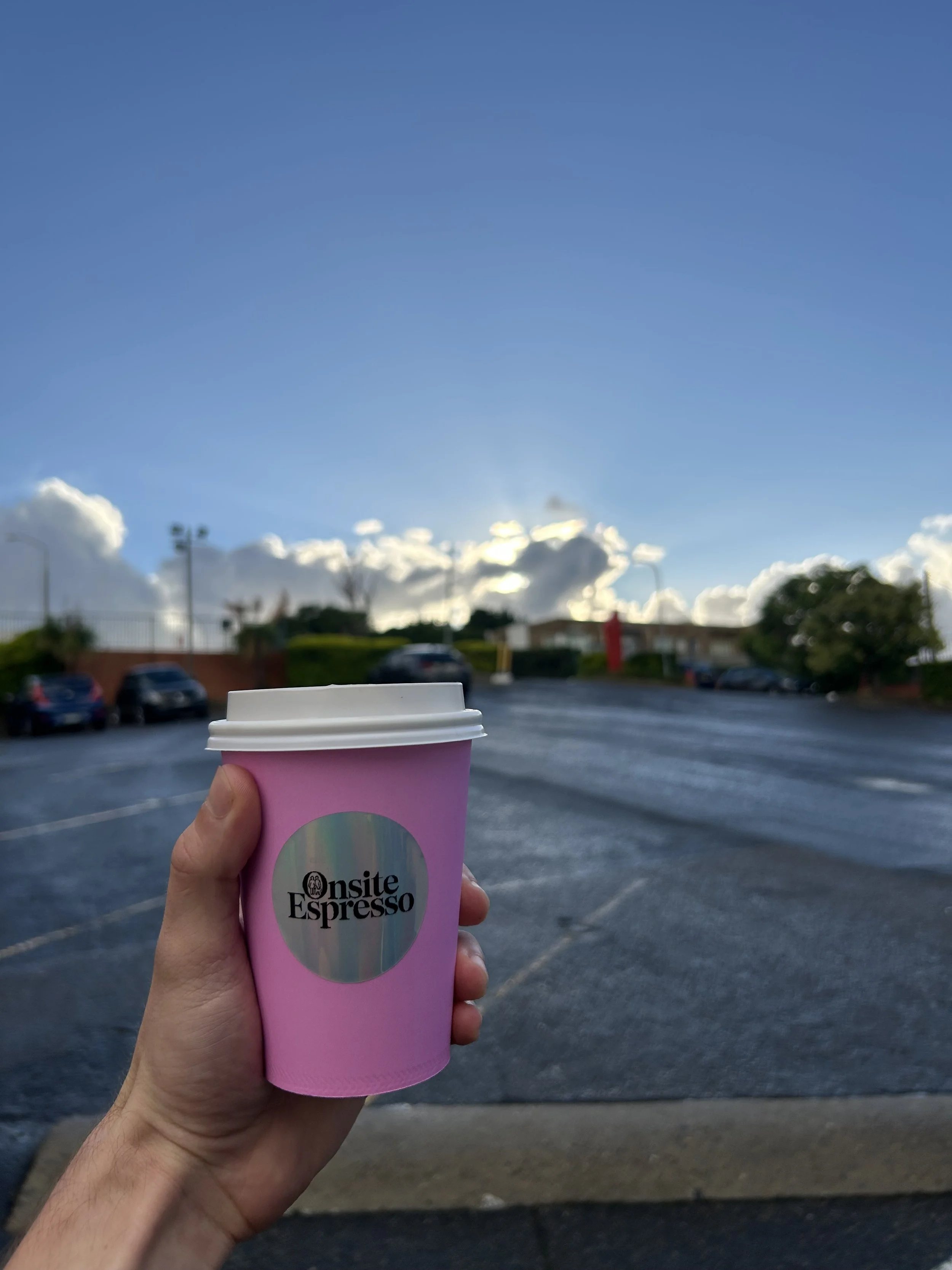 A person holding a pink paper coffee cup with a white lid in a parking lot during late afternoon or early evening, with clouds and blue sky in the background.