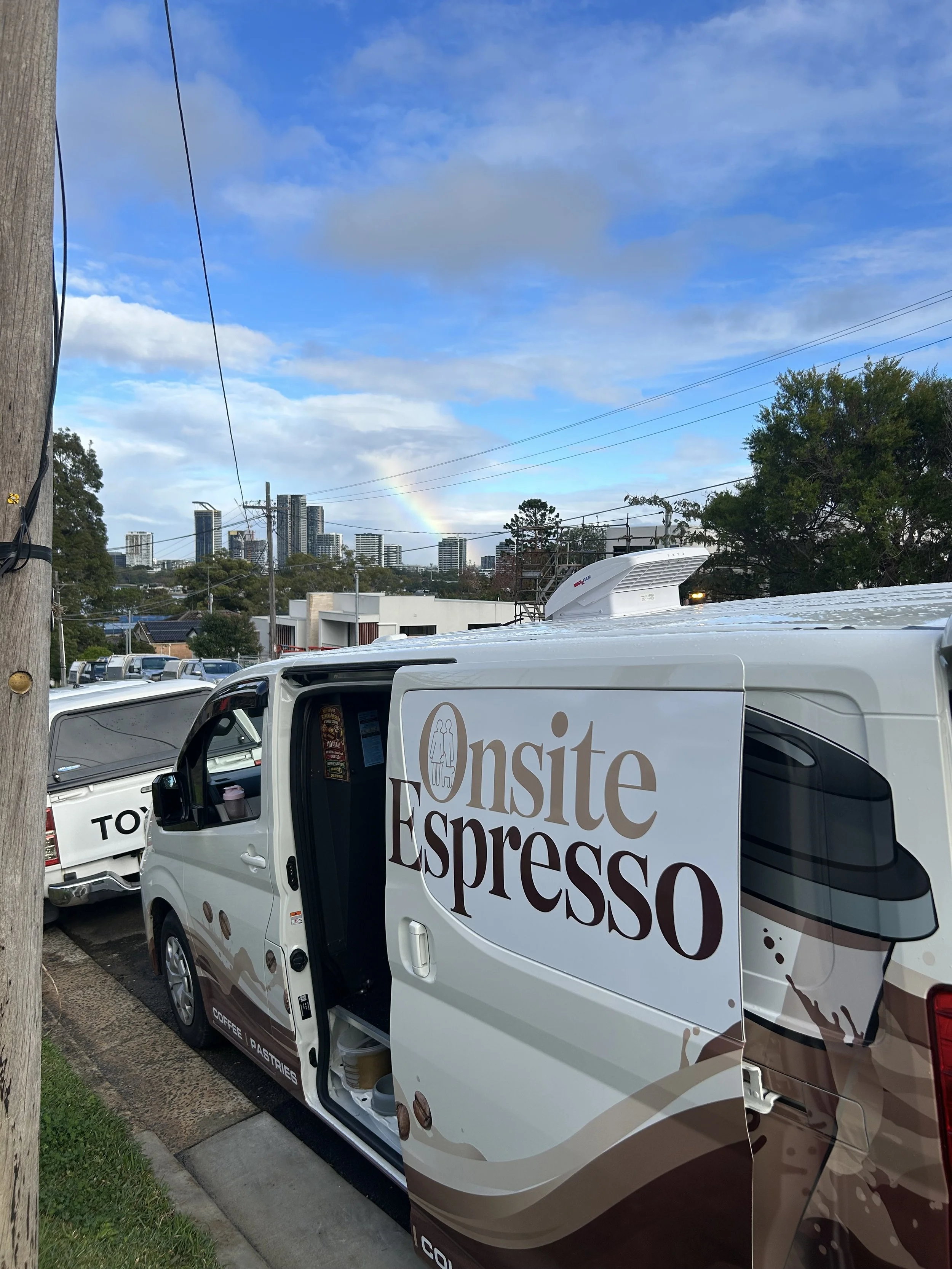 A white van with a sign that reads 'Onsite Espresso' parked on a street. In the background, there are city high-rise buildings, trees, utility poles, and a rainbow in the sky with clouds.