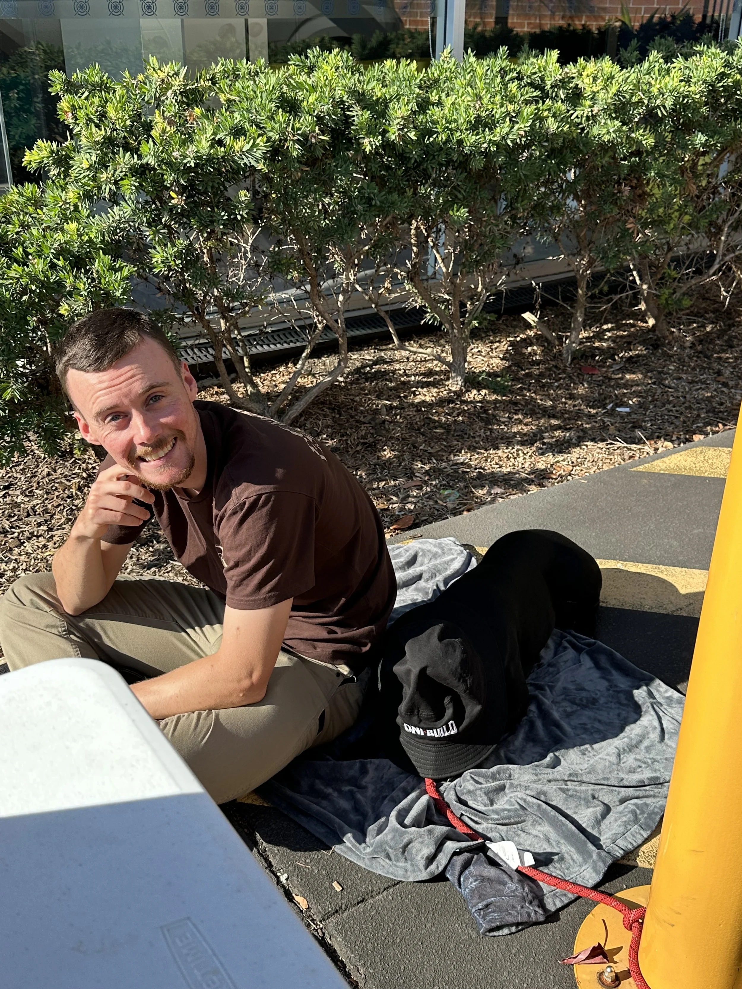 A man with short brown hair and a beard, wearing a brown t-shirt and khaki pants, sitting on the ground outdoors near a bush and a building, smiling at the camera. There is a black dog lying on a blanket or clothing nearby, and a yellow traffic cone 