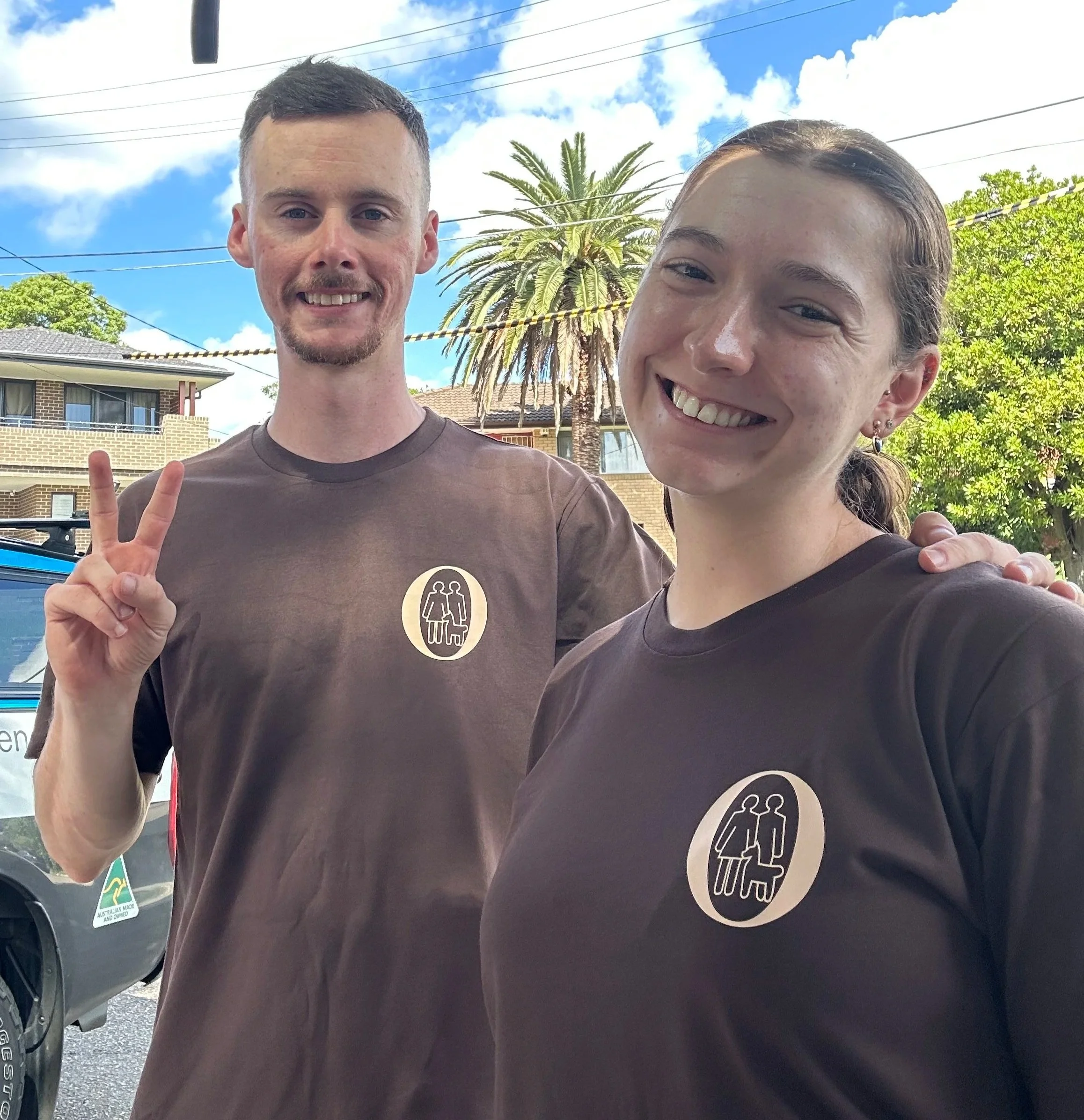 Two smiling people taking a selfie outdoors with a clear blue sky, a palm tree, and residential buildings in the background. The person on the left is making a peace sign with their hand.