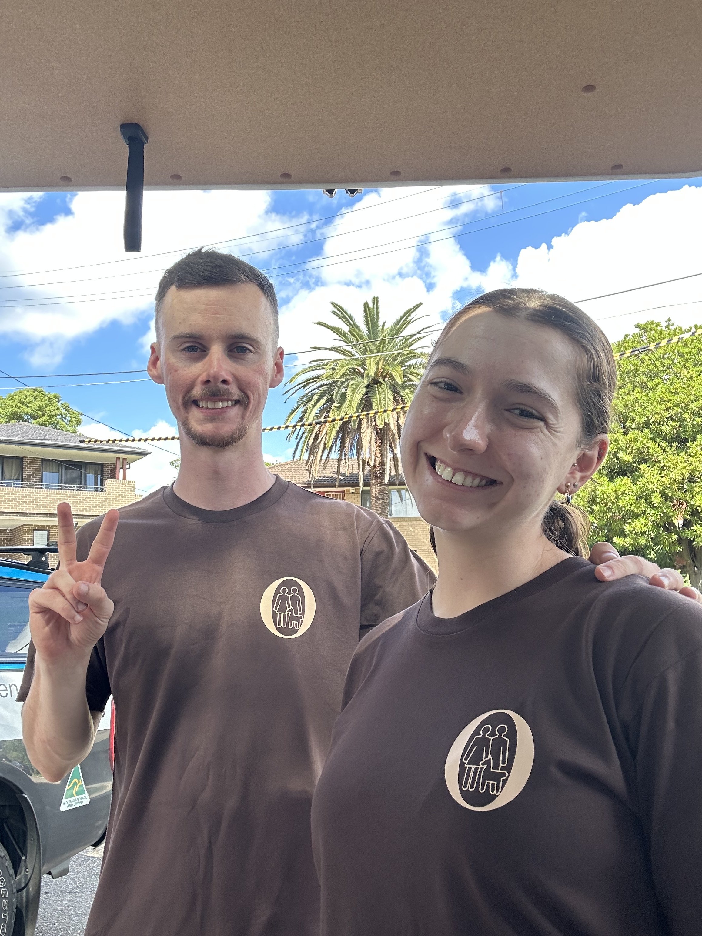 Two smiling young adults wearing matching brown T-shirts with a community logo, standing outdoors with blue sky, palm tree, and suburban houses in the background.