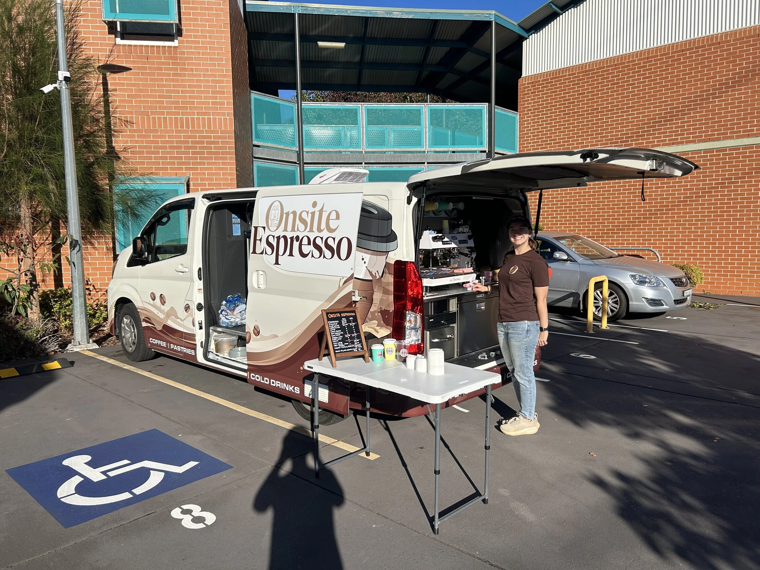 A coffee and pastry food truck named 'Onsite Espresso' parked in a parking lot next to a handicap parking space. A woman is standing in front of the truck smiling, and the truck has its back open showing the equipment inside. The background shows a b