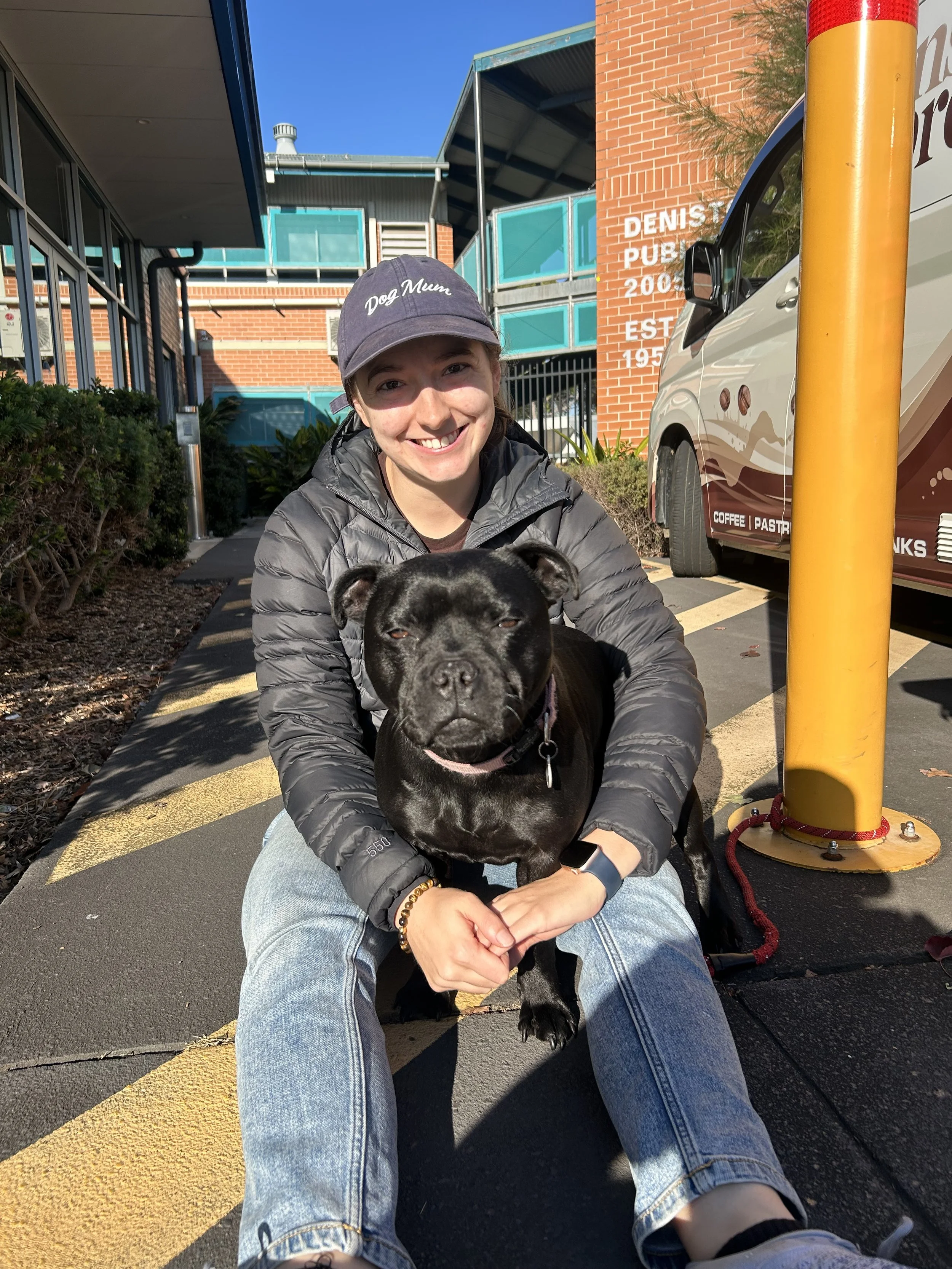 A woman sitting on the sidewalk holding a black dog, smiling at the camera, with a brick building, a parked vehicle, and a yellow pole in the background.
