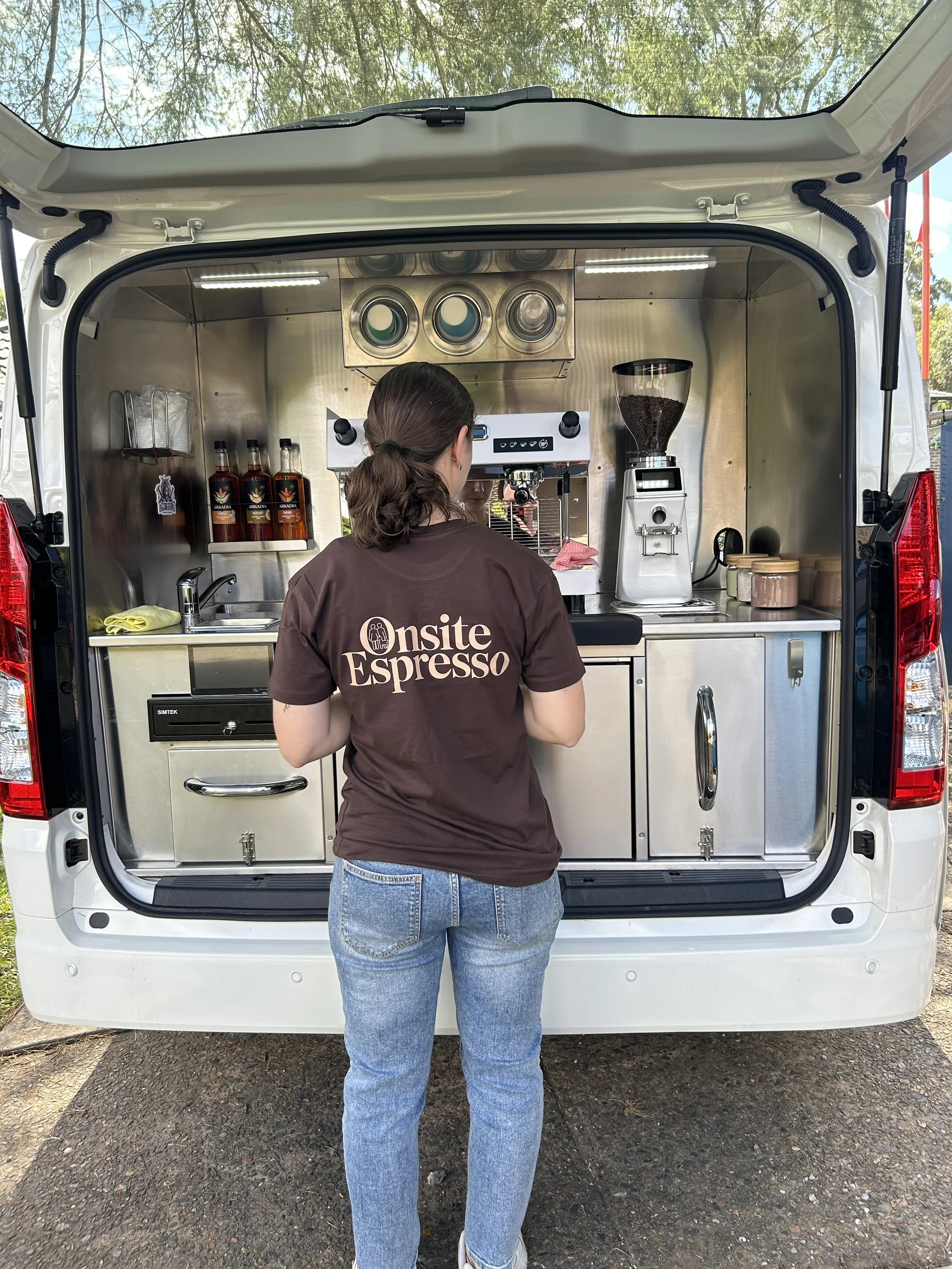 A woman with brown hair in a ponytail, wearing a brown 'Onsite Espresso' T-shirt and blue jeans, standing outside open the back of a white mobile coffee truck, with coffee bags, a coffee grinder, and other coffee-making equipment inside.