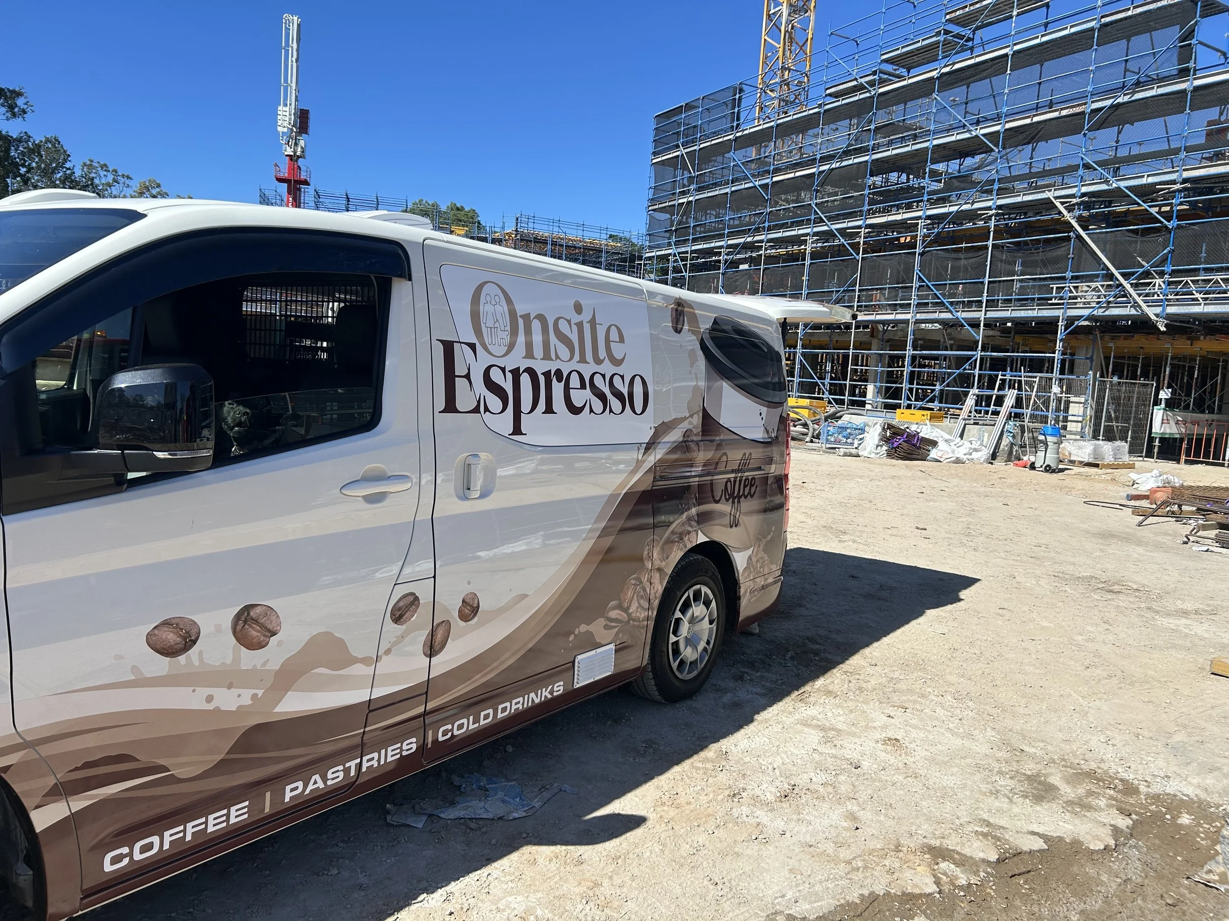 A white coffee van with a coffee bean graphic and the words "onsite Espresso" parked at a construction site with scaffolding and building framework in the background.