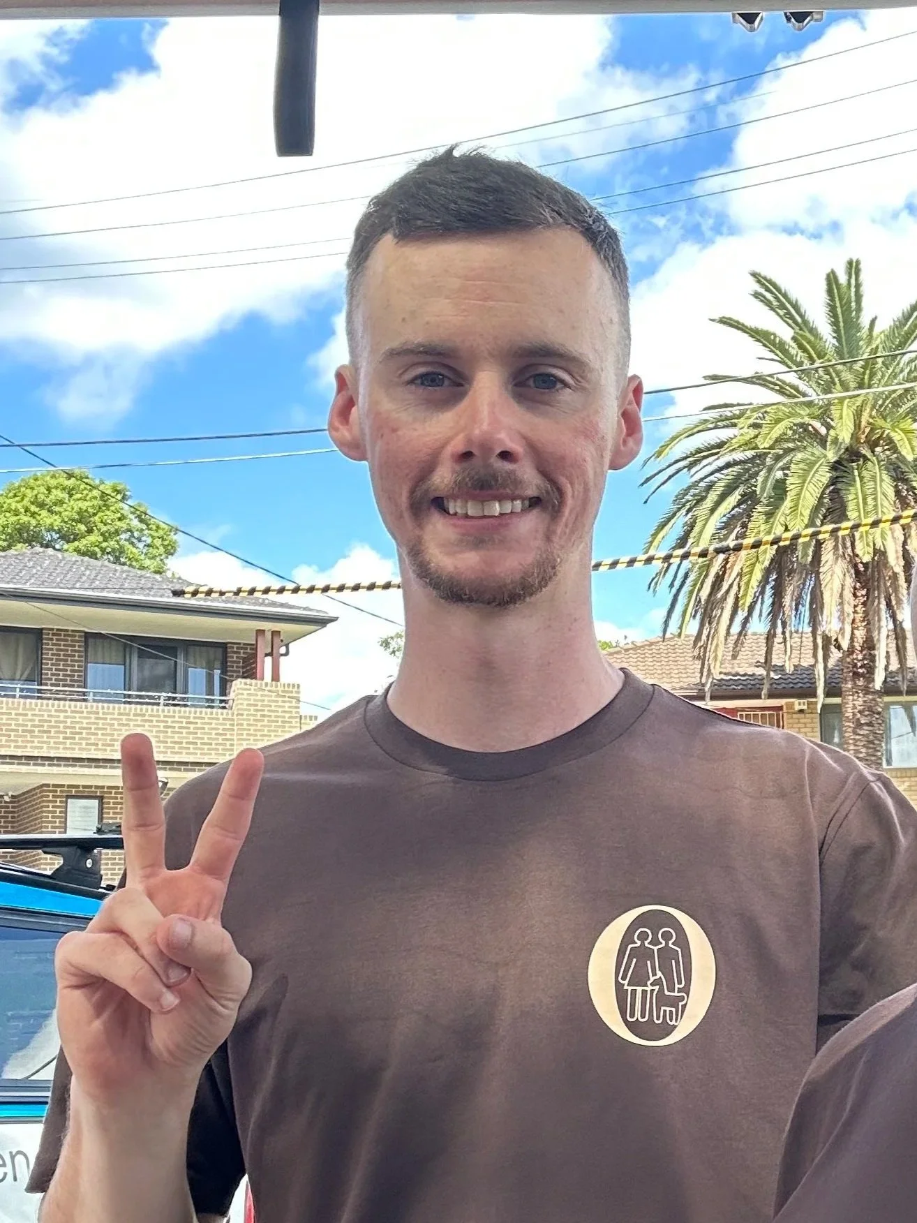 A man with short hair and a goatee making a peace sign with his hand outside, with a clear blue sky, palm trees, and residential buildings in the background.