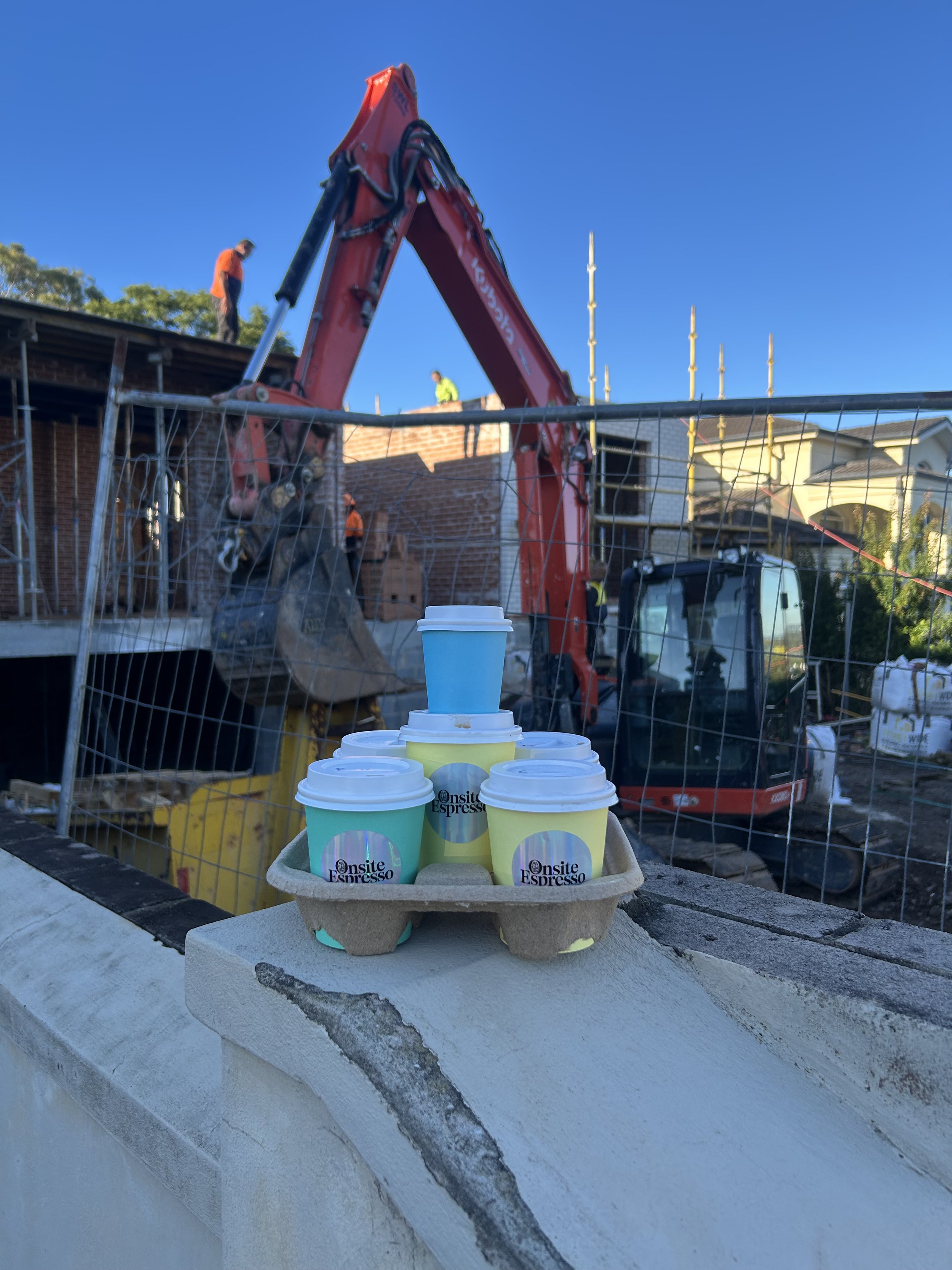 A tray with six cups of coffee labeled 'Onsite Espresso' placed on a ledge in front of a construction site. In the background, workers are visible working on a building with a red excavator and several houses under a clear blue sky.