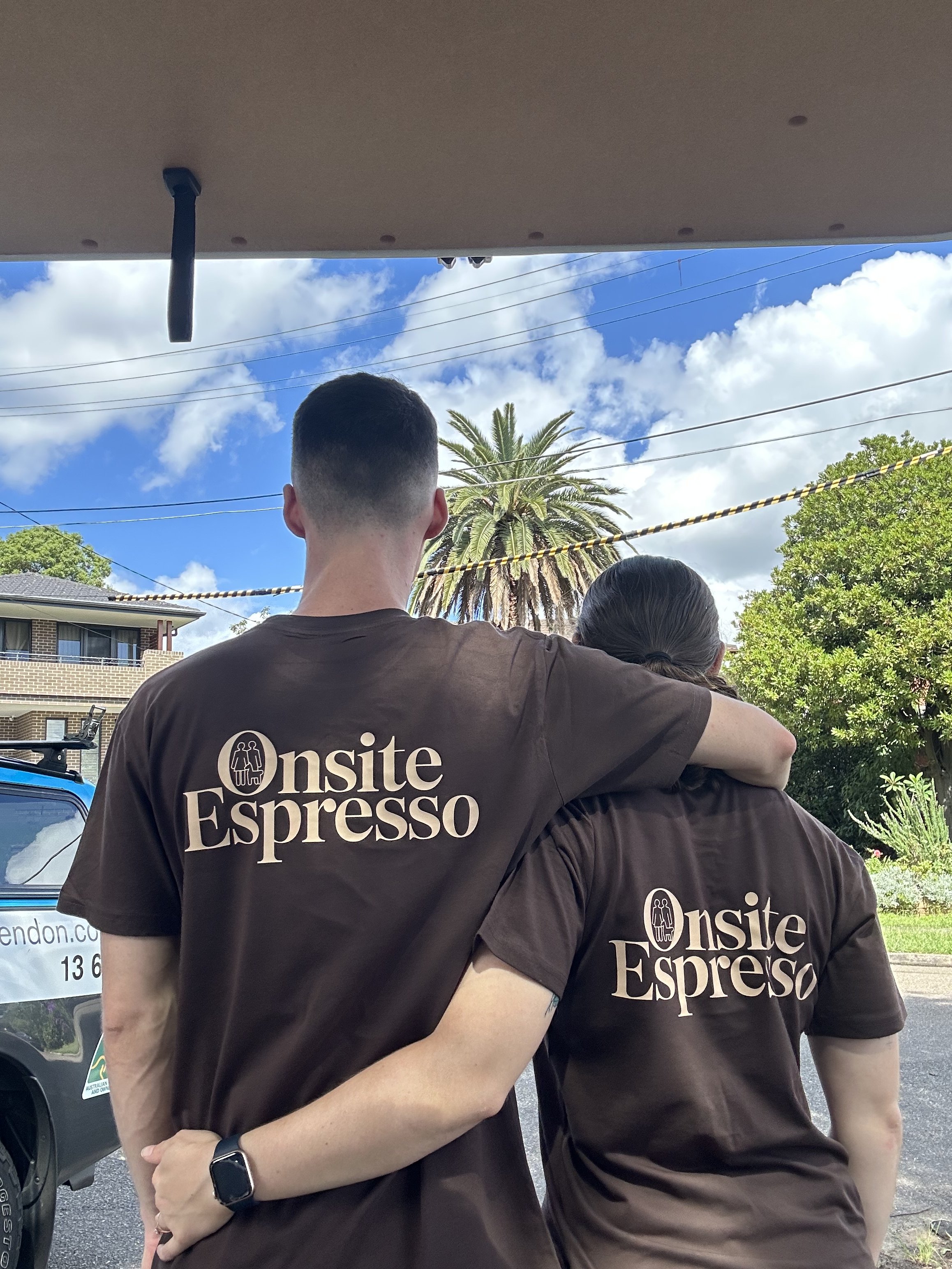 Two people wearing brown shirts with 'Onsite Espresso' logo, embracing outdoors, with a sunset, trees, and power lines in the background.