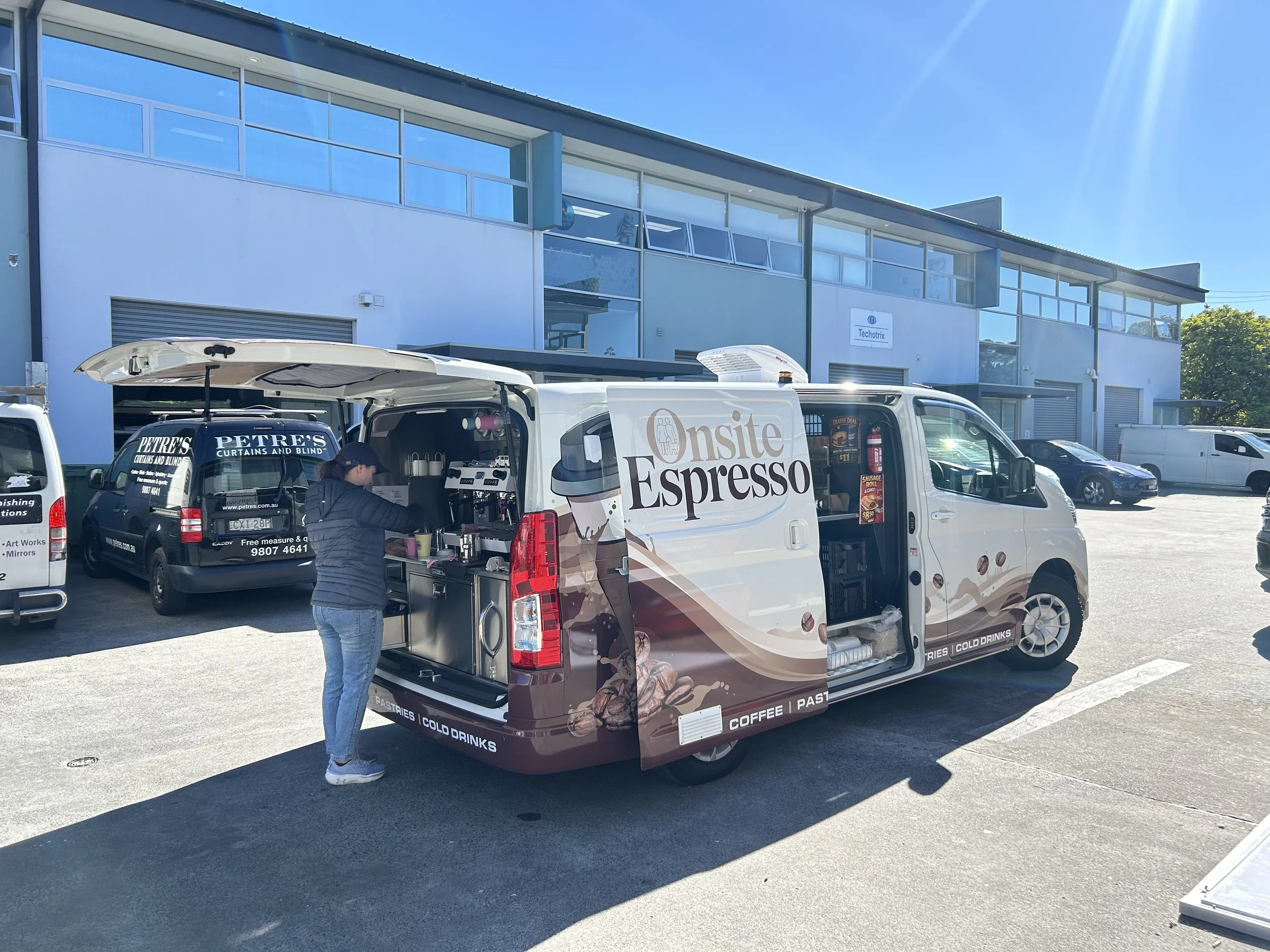 A white van with a coffee shop sign, open rear doors serving coffee and pastries, parked in a parking lot with cars and a modern building in the background.