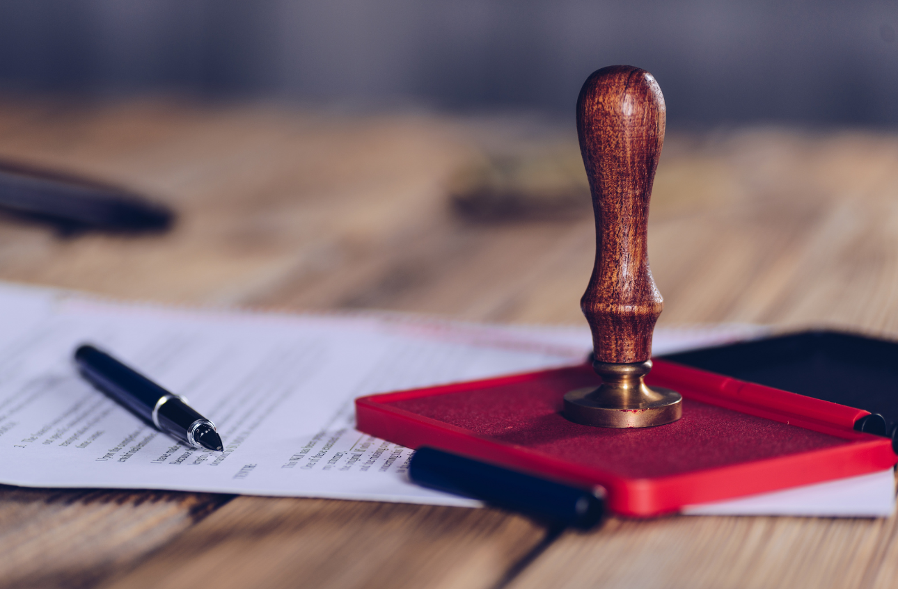 A rubber stamp with a wooden handle resting on a red ink pad, which is placed on documents and surrounded by pens on a wooden surface.