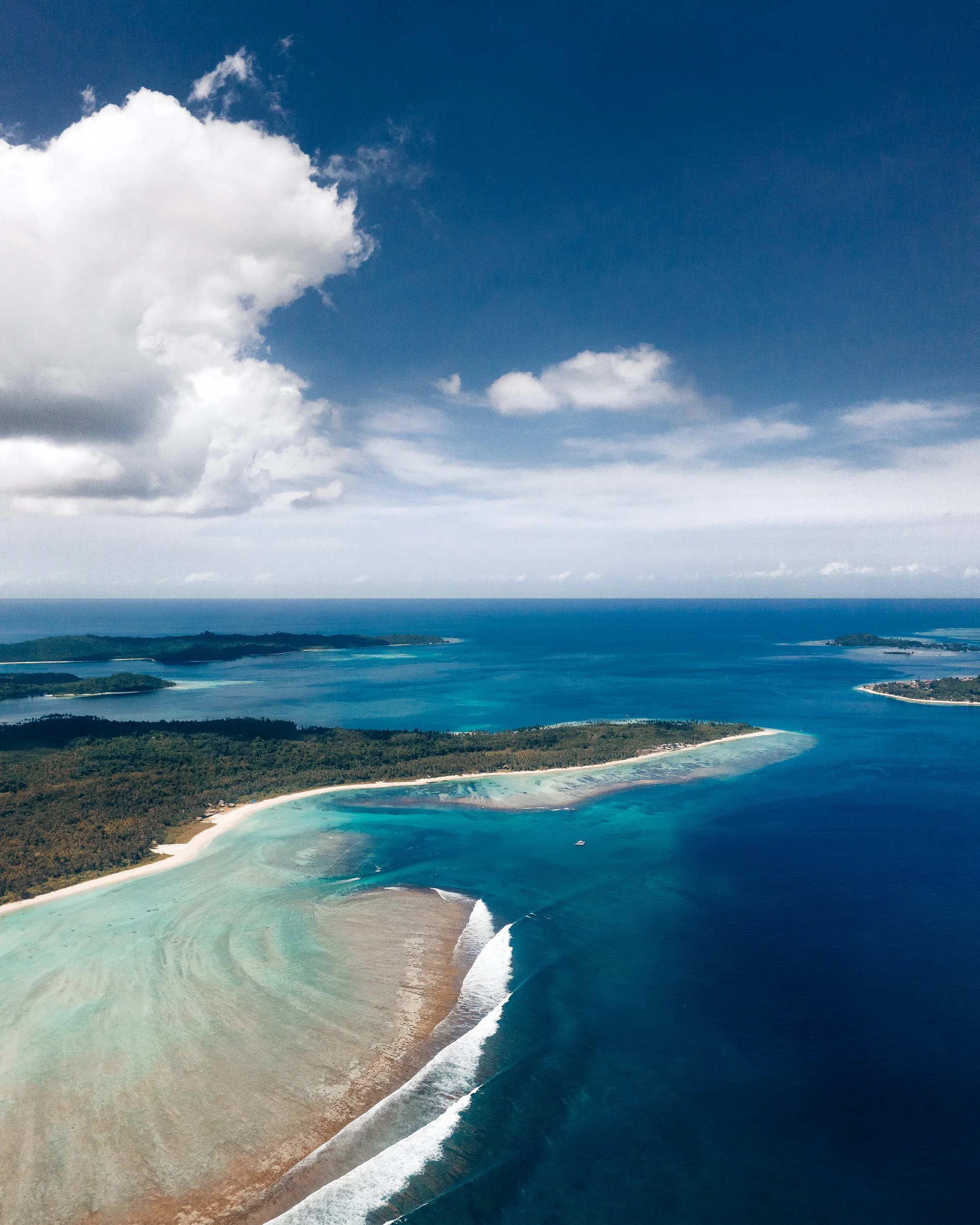 Aerial view of a coastline with blue ocean, sandy beaches, and lush green forests under a partly cloudy sky.
