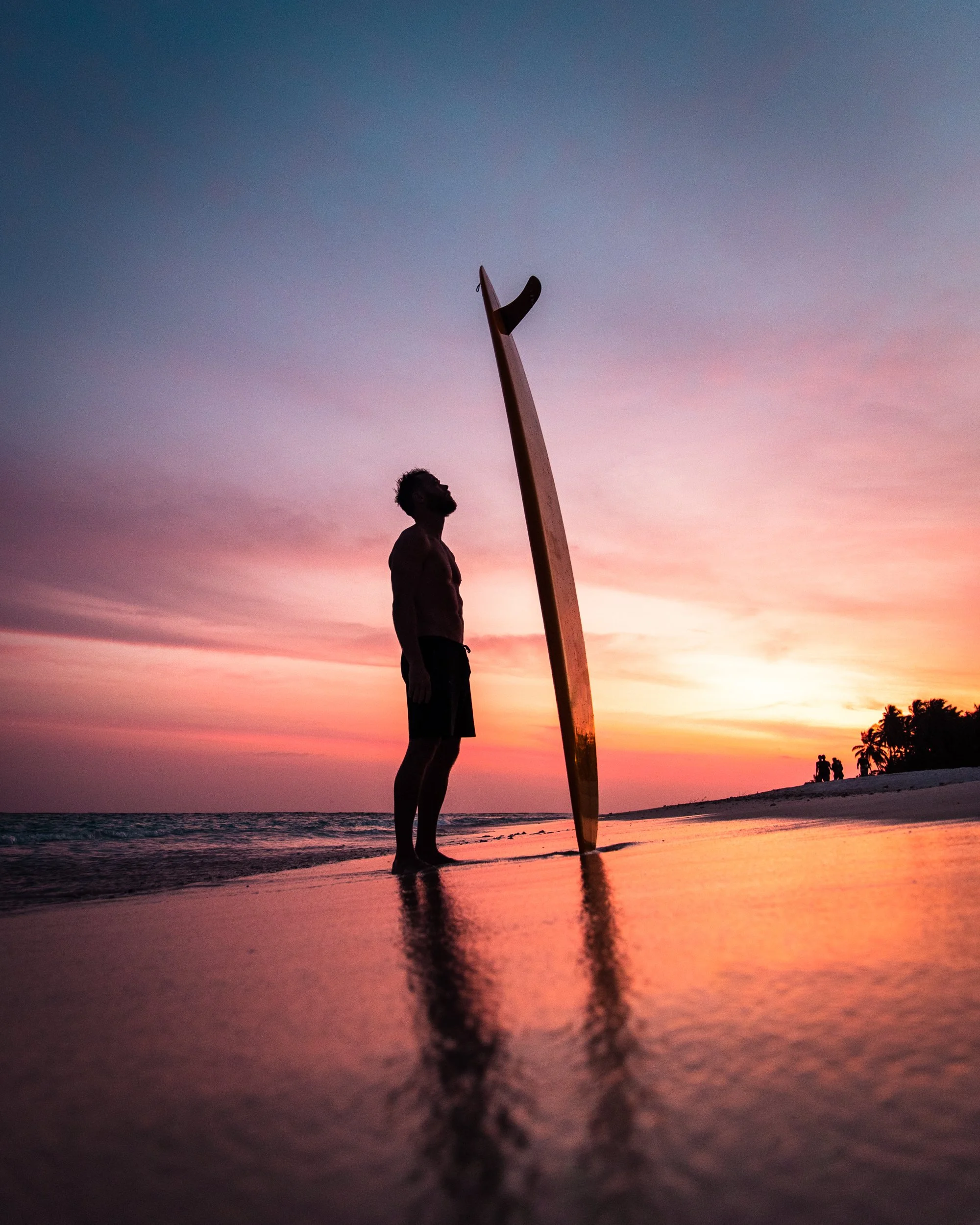 Silhouette of a man standing on the beach near the ocean at sunset, with a surfboard upright in the sand, and palm trees in the distance.