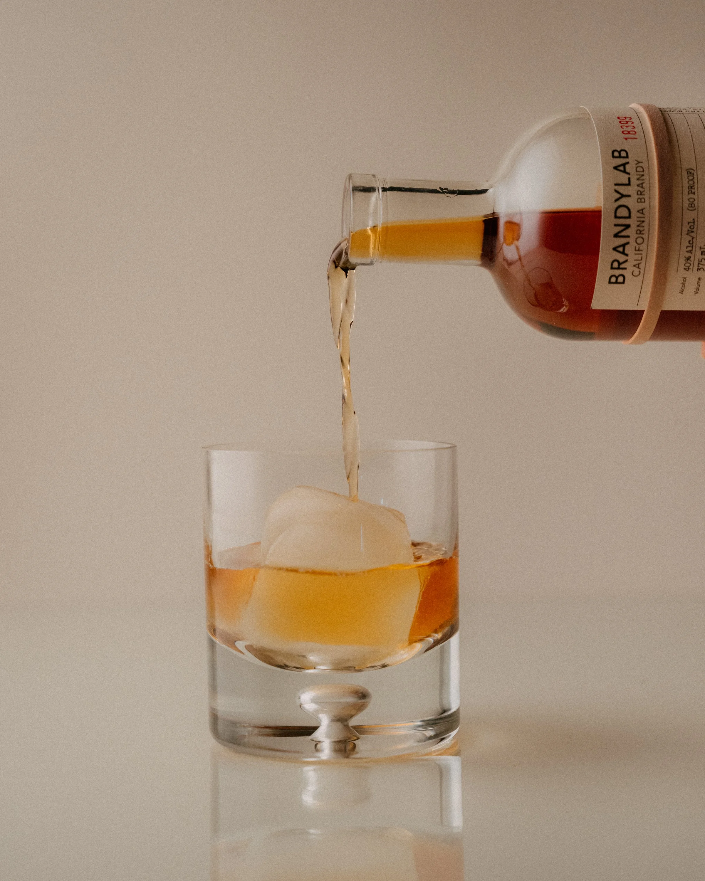 A glass with a drink and ice cubes, with a pour of amber-colored liquor from a bottle labeled 'Brandylab California Brandy'.