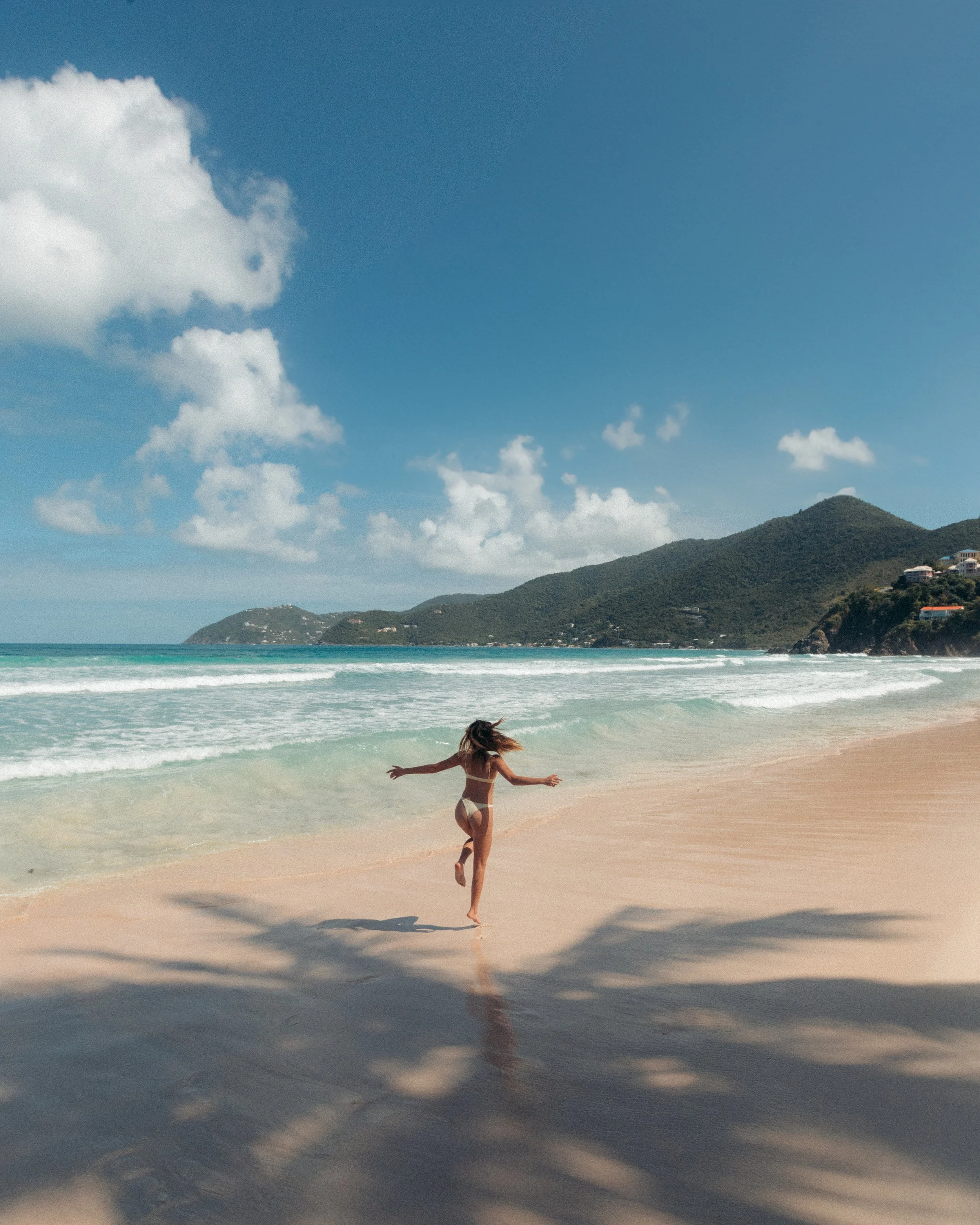 A young woman in a white bikini running along the beach shoreline with her arms outstretched. The ocean waves are crashing on the sand, and a green, hilly landscape can be seen in the distance under a partly cloudy sky.