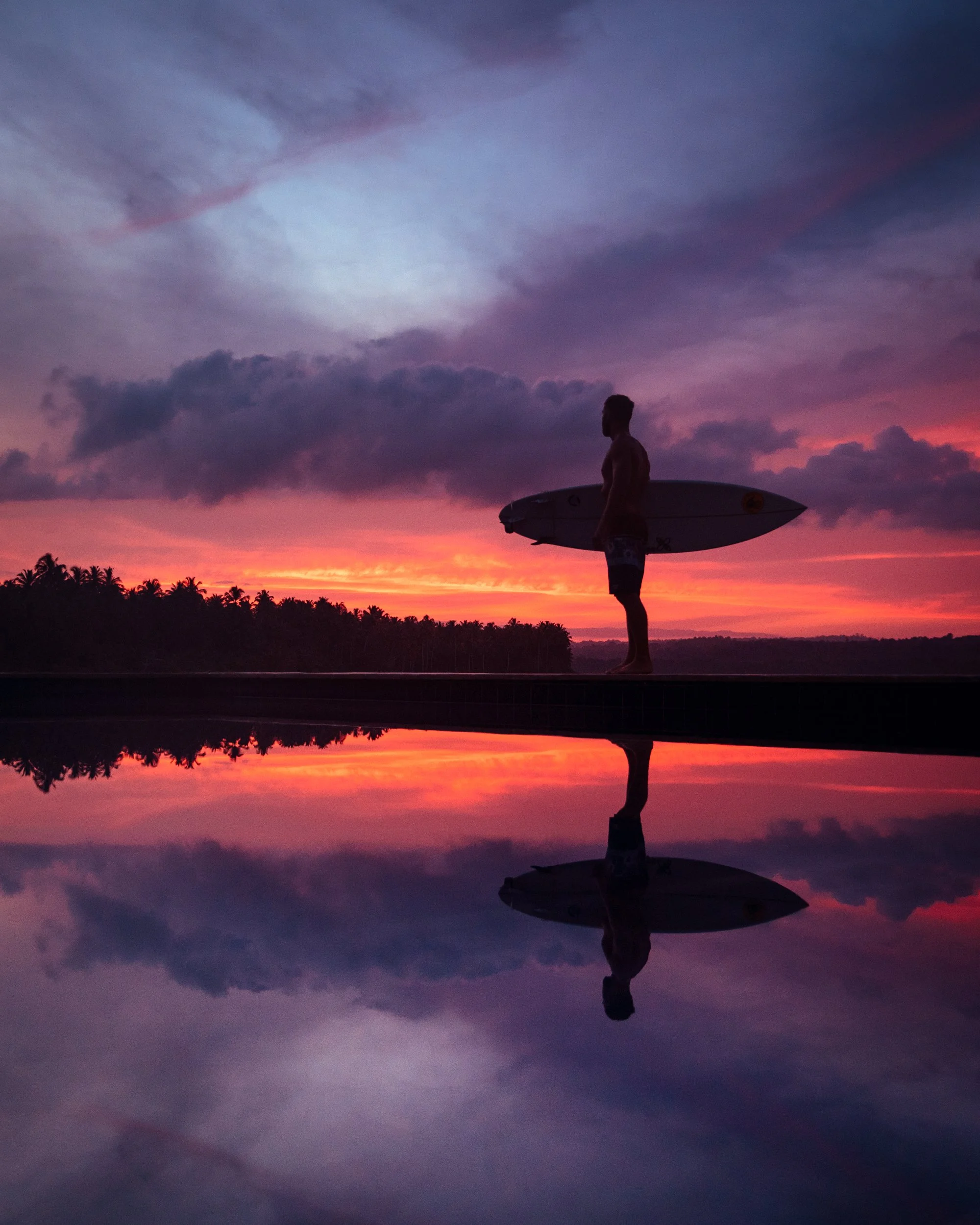 Silhouette of a man holding a surfboard at sunset with colorful clouds and a reflected sky on water.